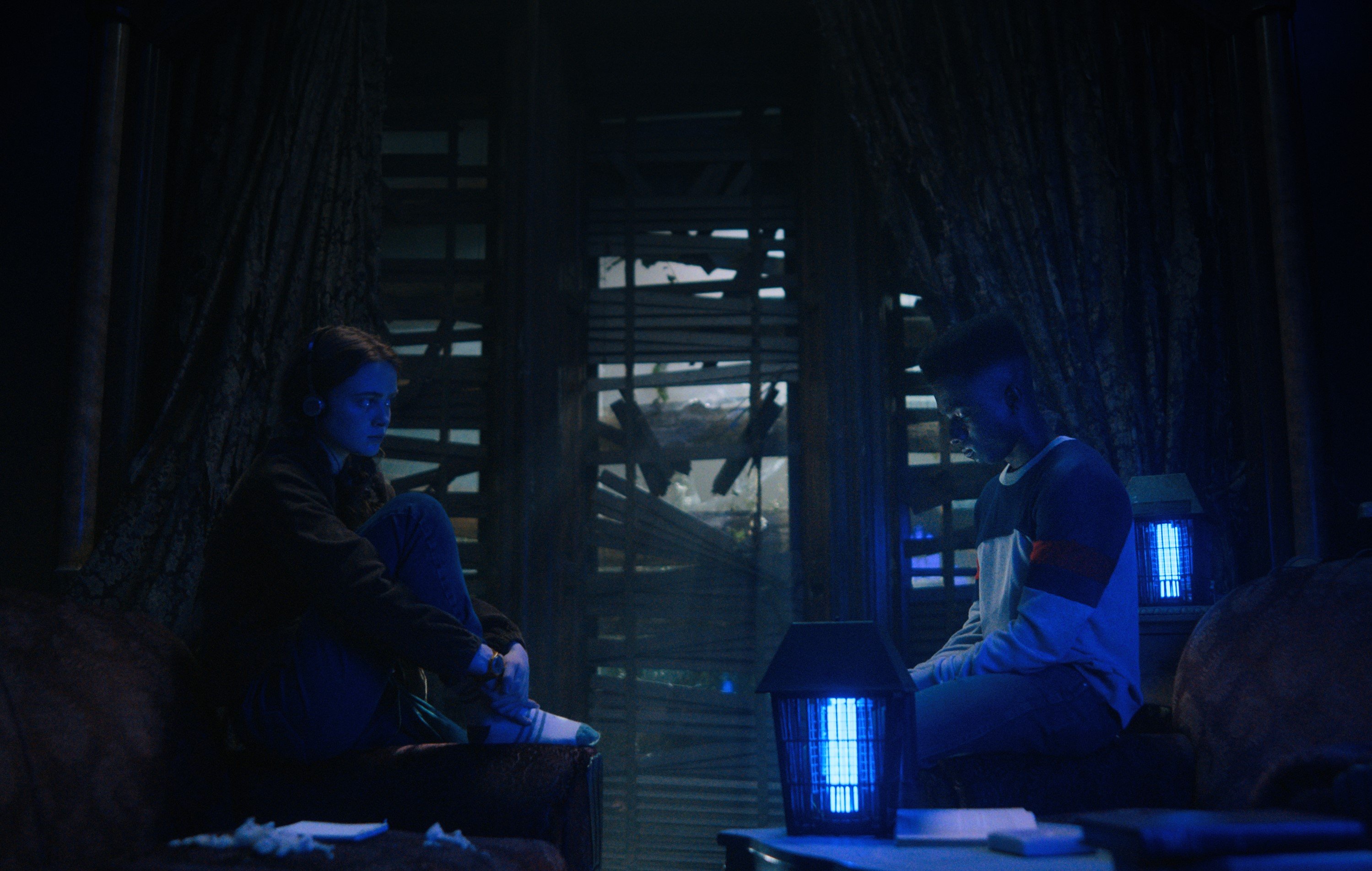 Max and Lucas in 'Stranger Things' Season 4, the latter of whom reads a book at the end that teases the Duffer Brothers' new TV show. In this photo, the pair is sitting in a dark room with blue lighting and facing one another.