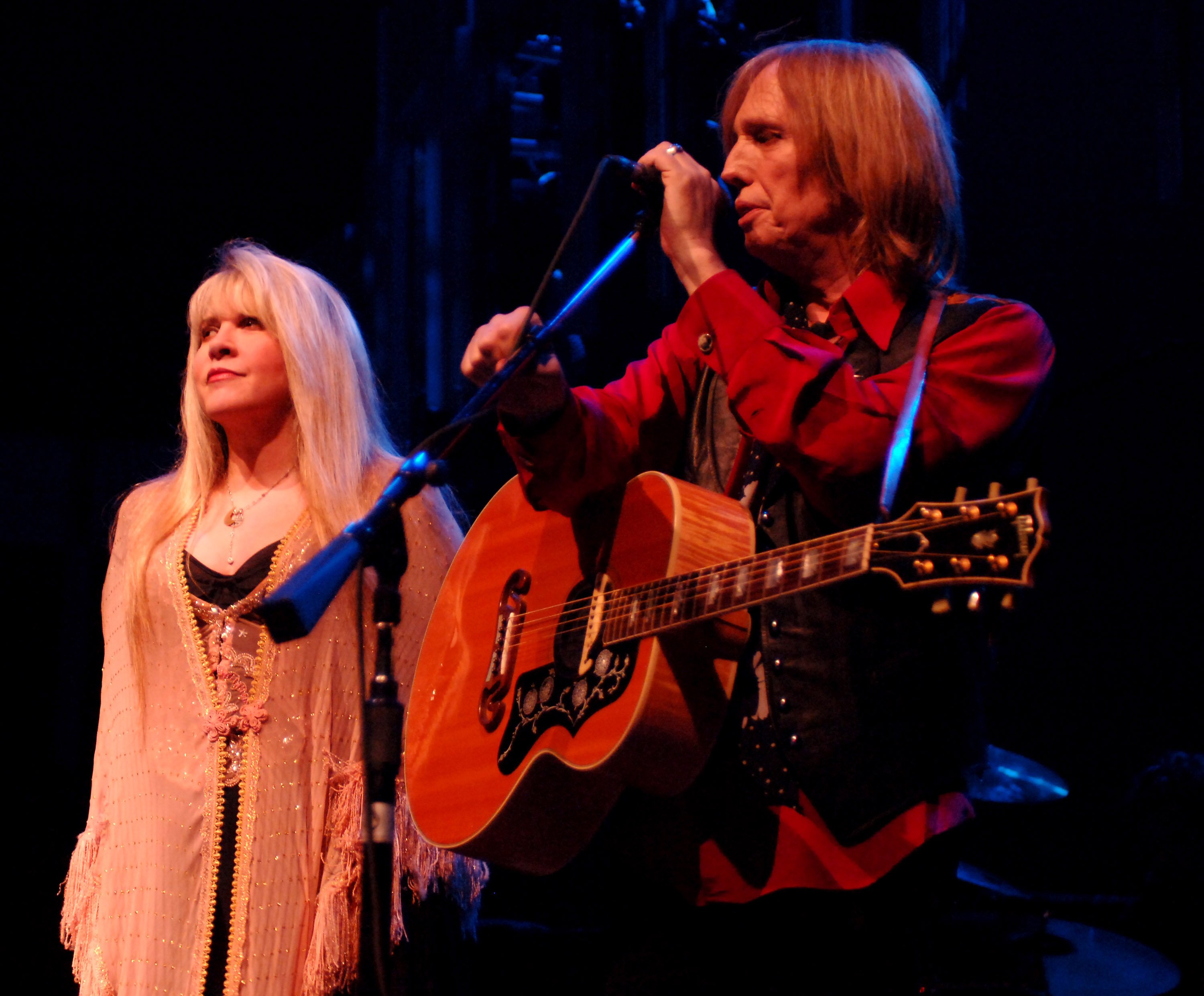 Stevie Nicks stands next to Tom Petty who has a guitar and adjusts a microphone.