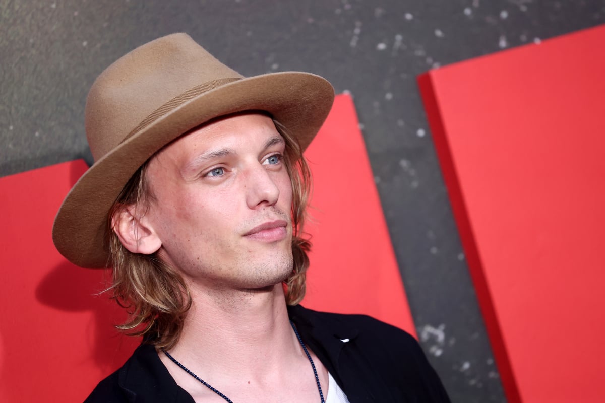 Stranger Things star Jamie Campbell Bower wears a tan hat, black sweater, and white shirt.