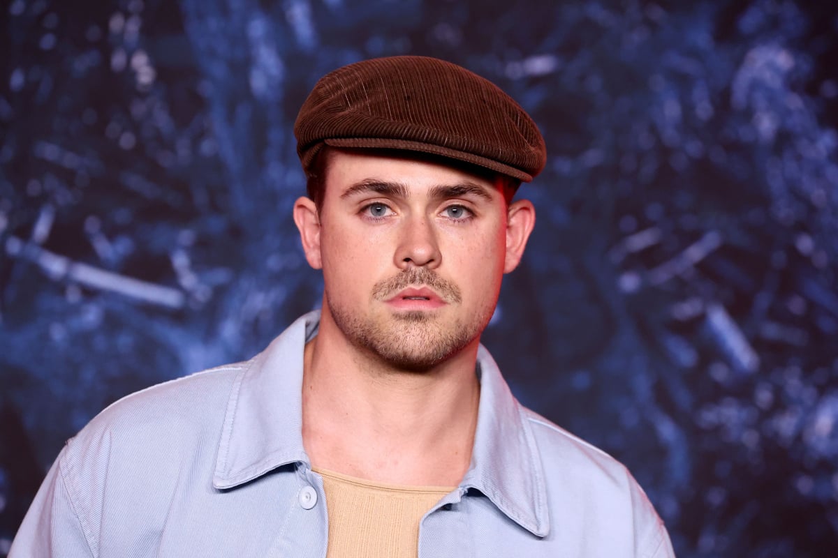 Dacre Montgomery attends Netflix's Stranger Things Season 4 Premiere at Netflix Brooklyn. Montgomery wears a newsboy hat and a tan shirt with a blue button up over it. 