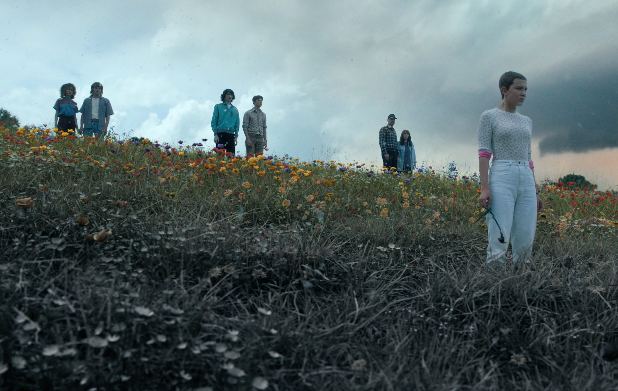 The 'Stranger Things' cast, who will return when the Duffer Brothers start filming season 5. They're standing in a field that's half green and half grey and looking out at something off-screen.