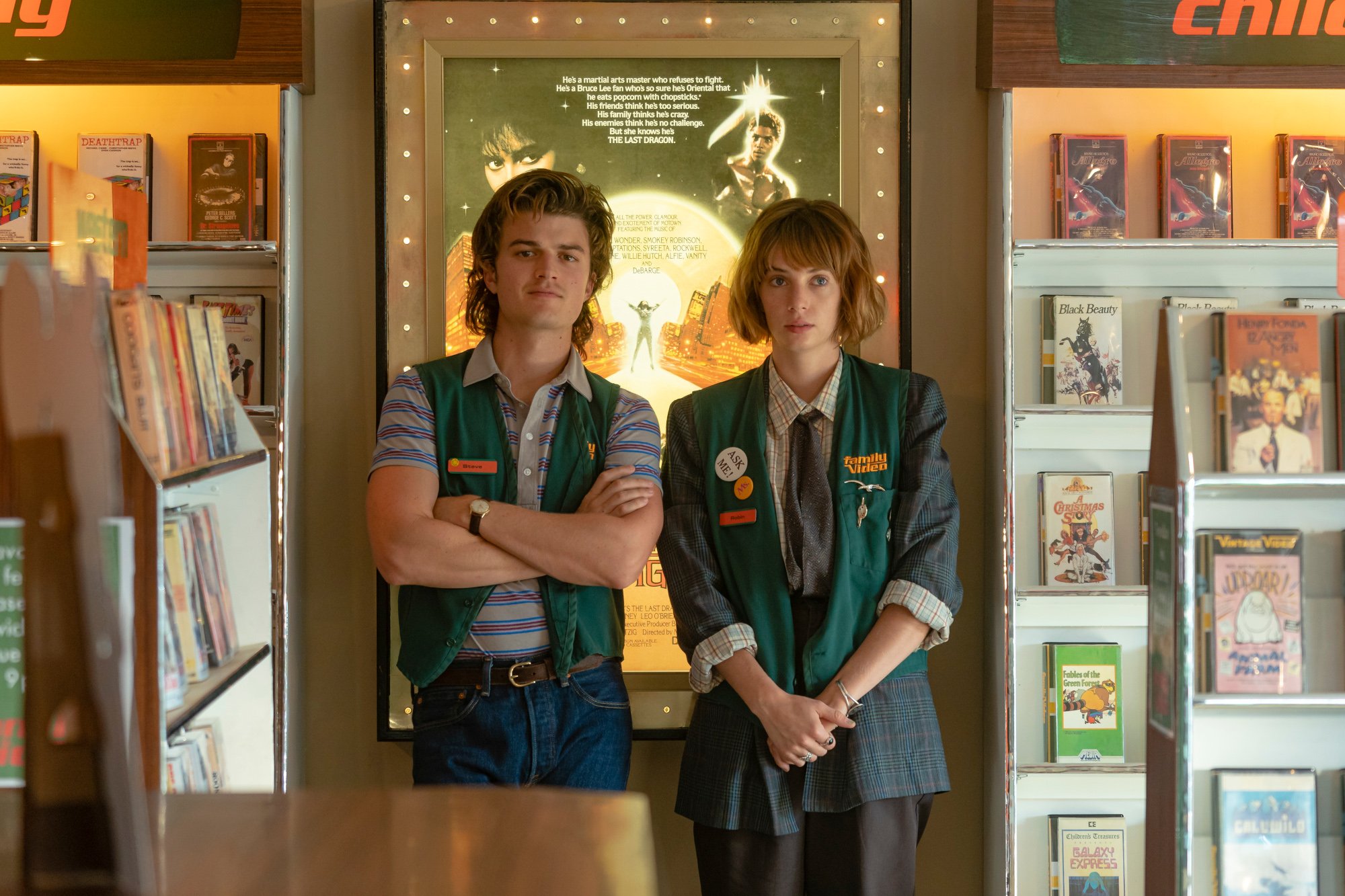 Joe Keery as Steve Harrington and Maya Hawke as Robin Buckley in 'Stranger Things,' which is among the TV shows and movies to watch over the Fourth of July weekend. The pair is standing next to one another in a video store. They're both wearing green vests.