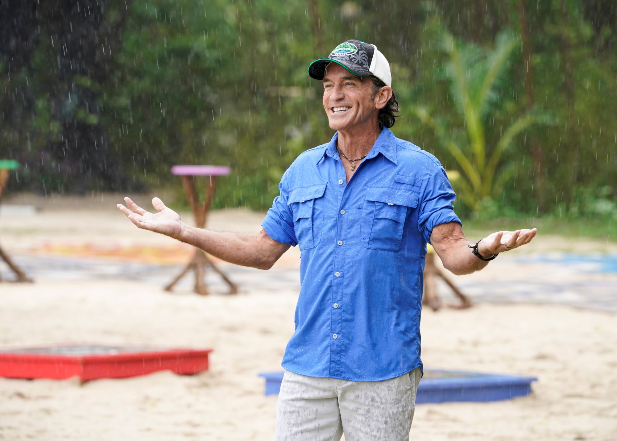 'Survivor' Season 43 host Jeff Probst wears a light blue shirt, tan pants, and a black, white, and green 'Survivor' baseball cap while standing in the rain.