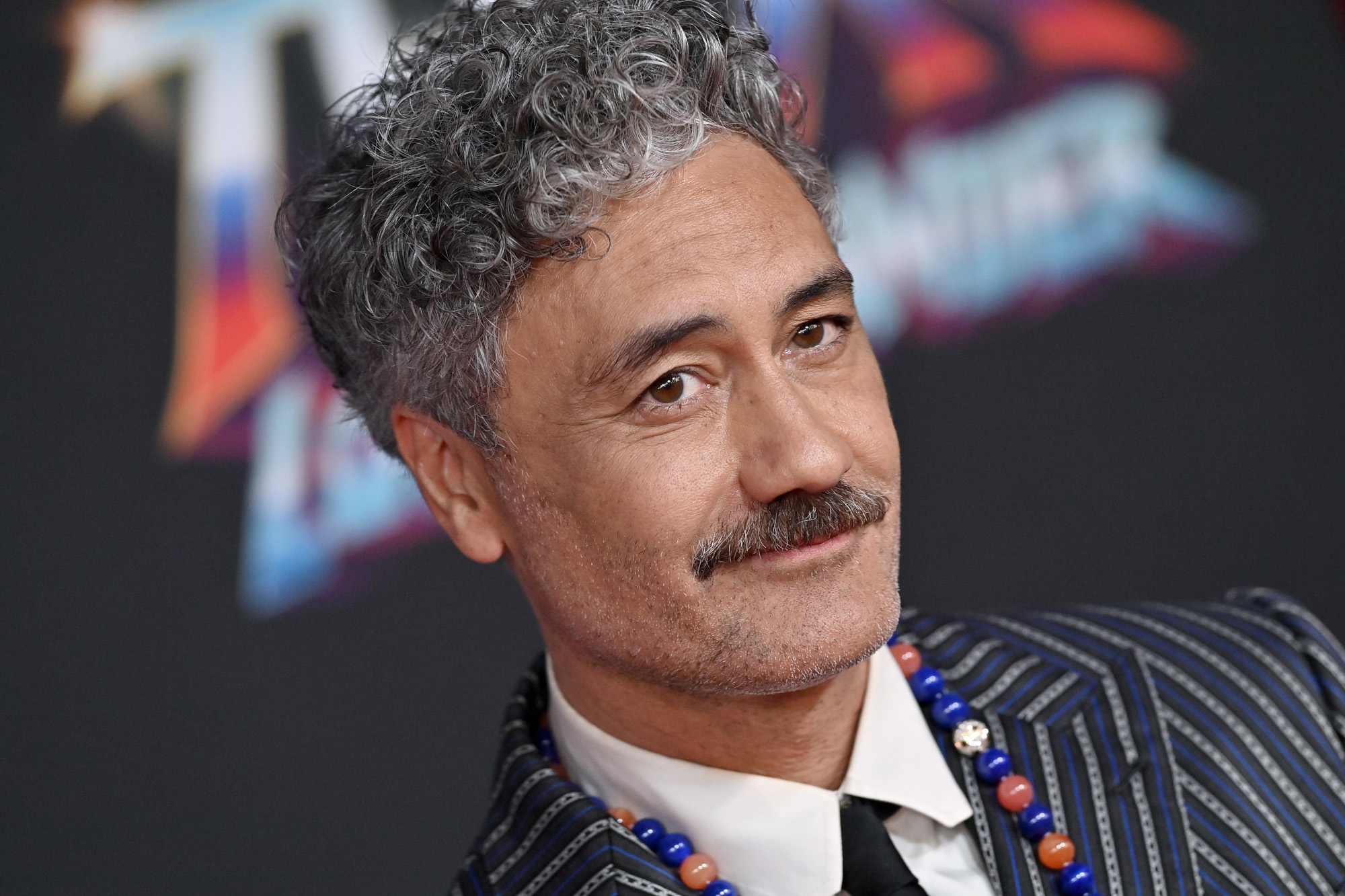 Taika Waititi, who was seen wearing a blurred-out 'One Piece' shirt, suggesting he is a fan of the anime. In this photo, he is wearing a blue, white, and black suit and smiling.