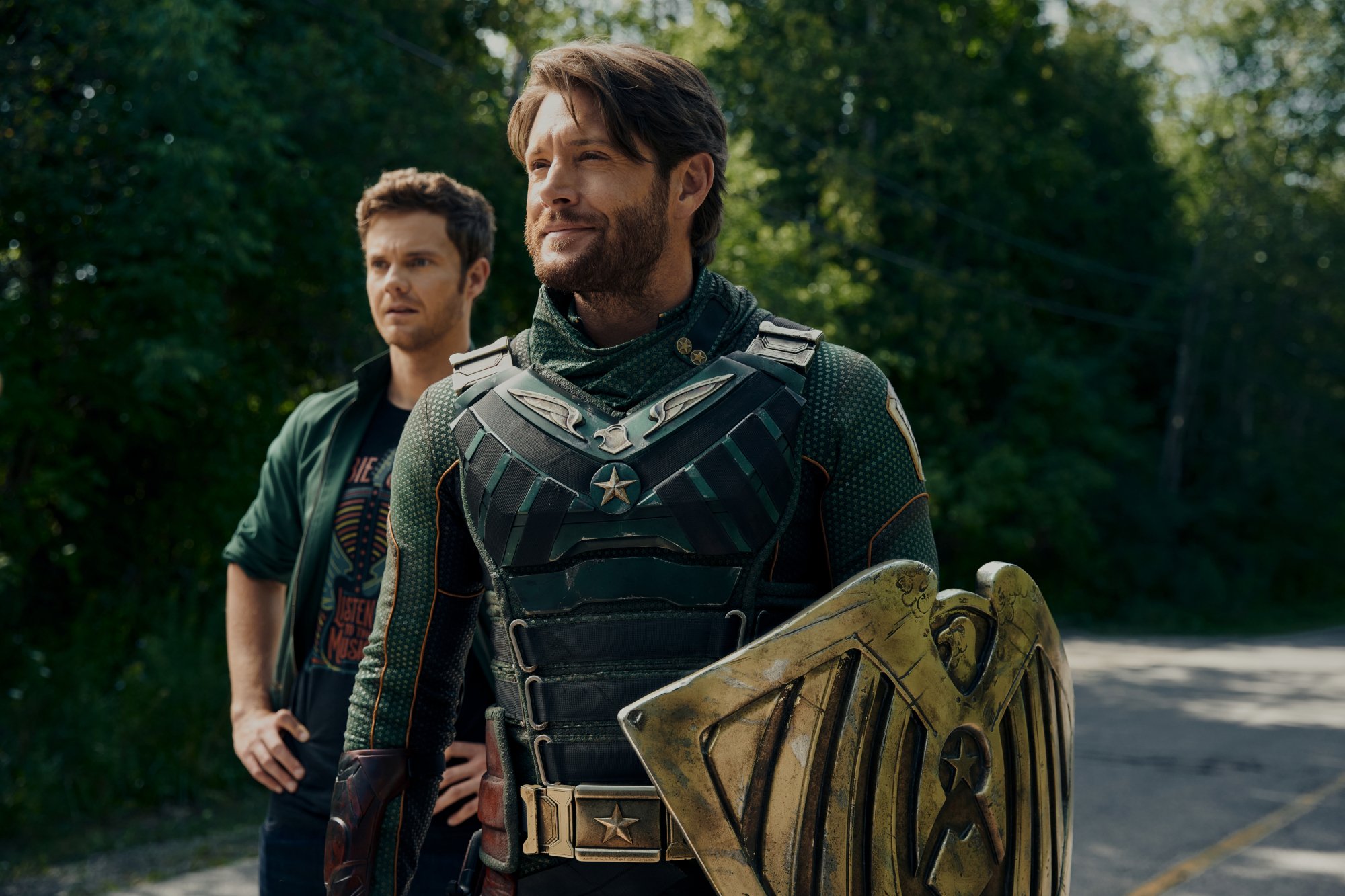 Jack Quaid and Jensen Ackles as Hughie Campbell and Soldier Boy in 'The Boys' Season 3, which has an episode 8 (finale) release date of July 8. Soldier Boy is wearing a green and black suit and holding a shield. Hughie is standing slightly behind him.