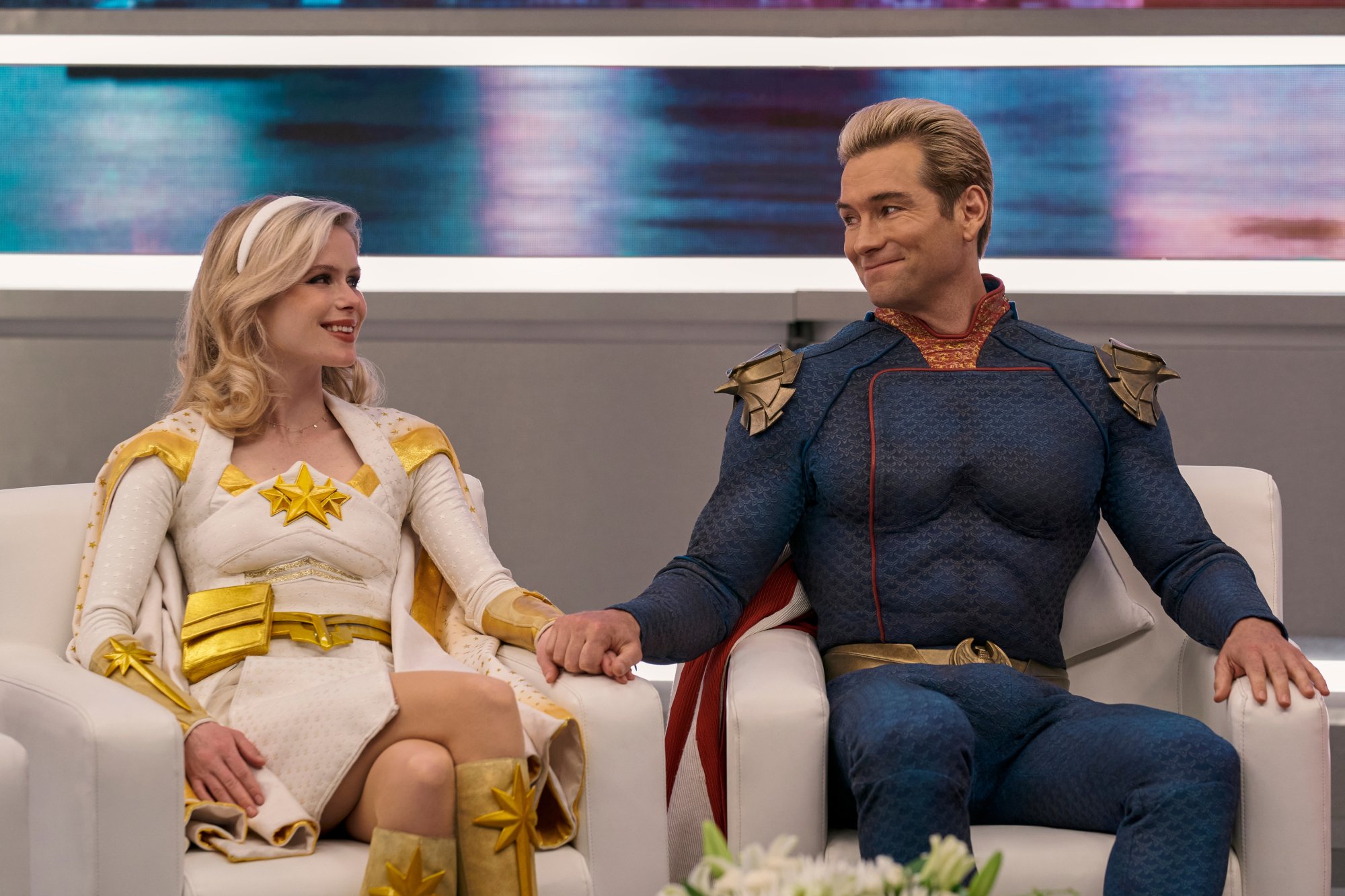 Erin Moriarty and Antony Starr as Starlight and Homelander in 'The Boys,' which will get a spinoff series ahead of season 4. The two are sitting next to one another in white armchairs and smiling. Homelander is holding Starlight's knee.