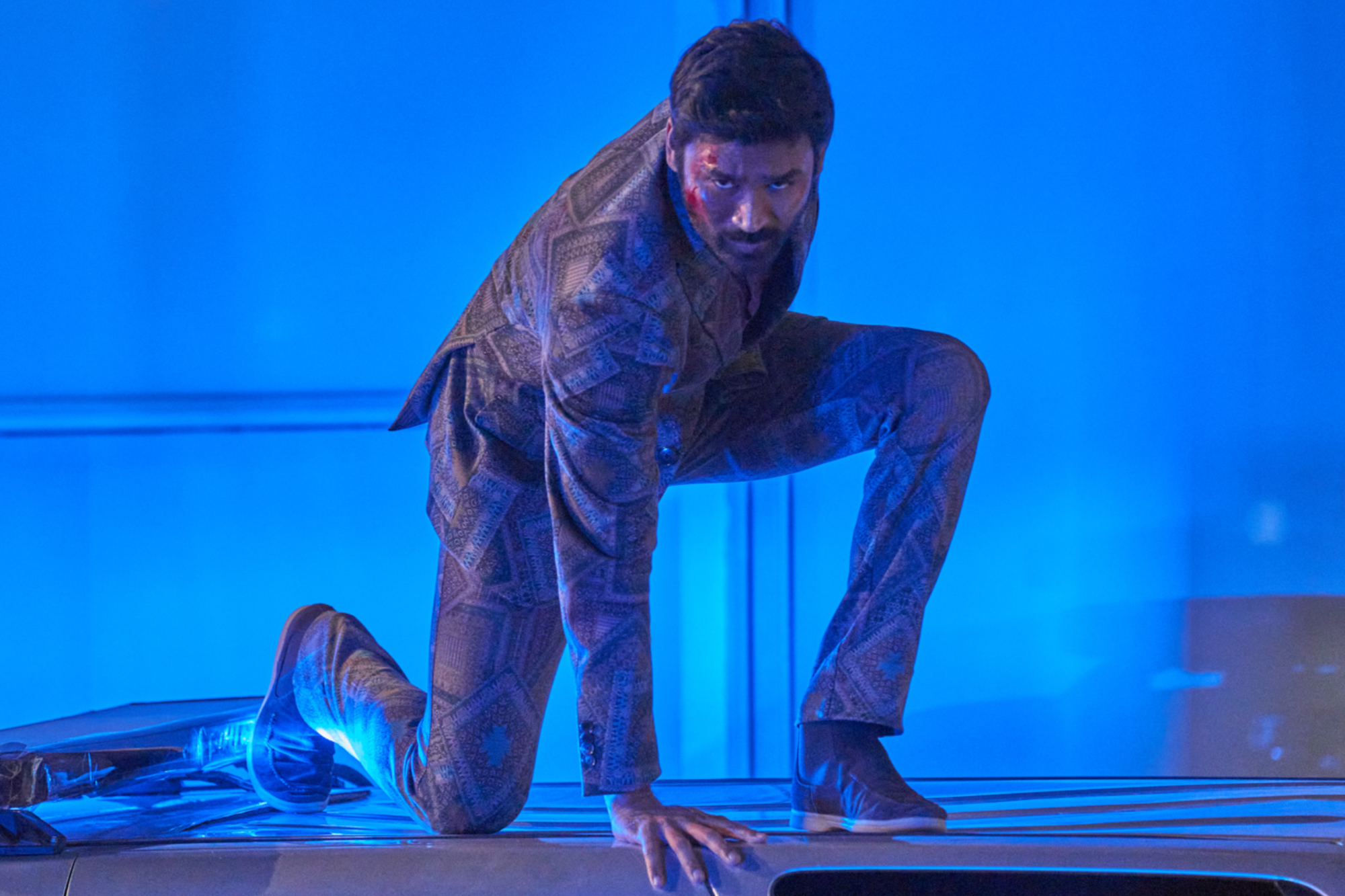 'The Gray Man' Dhanush as Avik San in an action pose with his hand on the top of a car wearing a patterned suit and some blood on his face