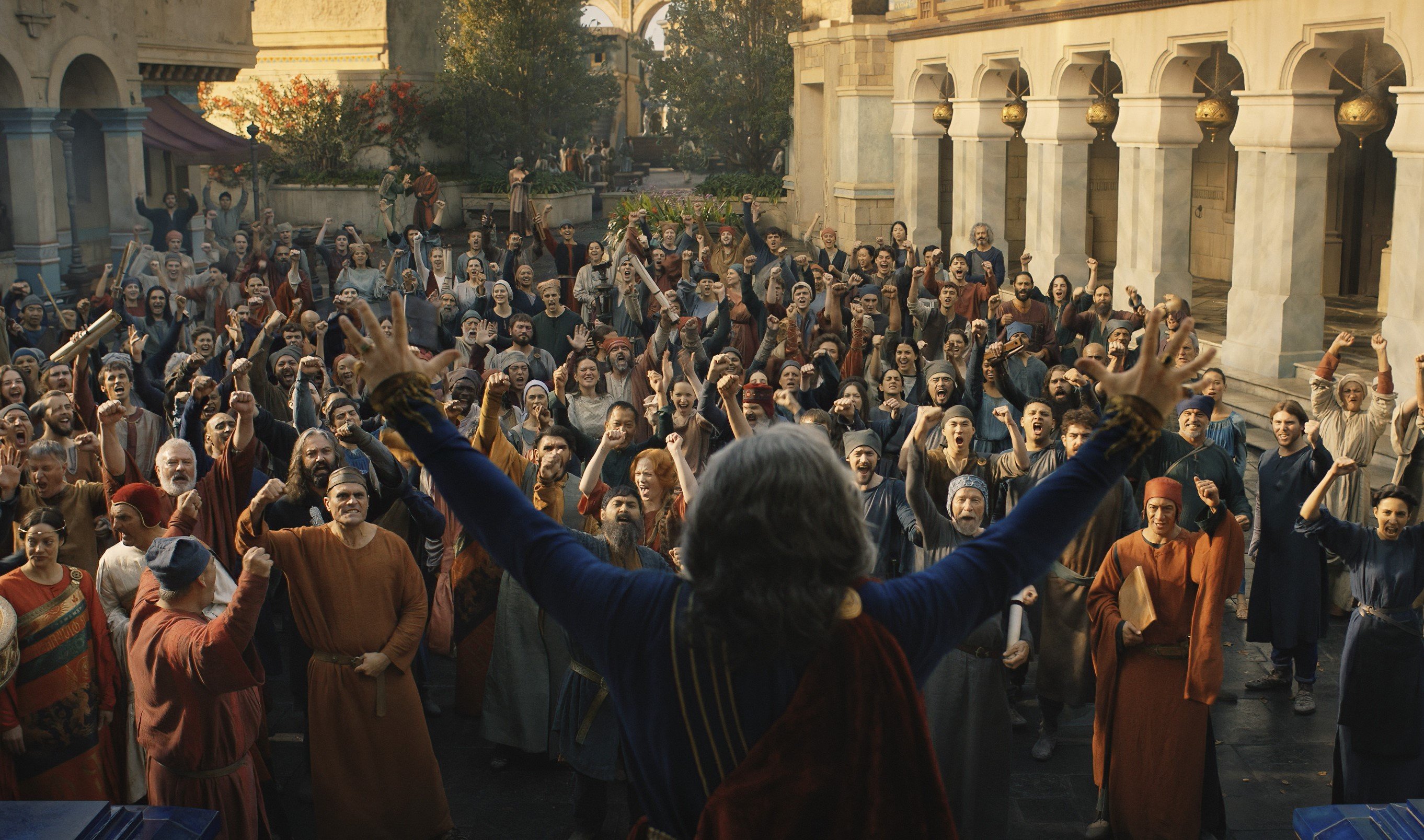 An image from 'The Lord of the Rings: The Rings of Power,' which is not adapting a single book. The image shows a man standing in front of a crowd and raising his arms.