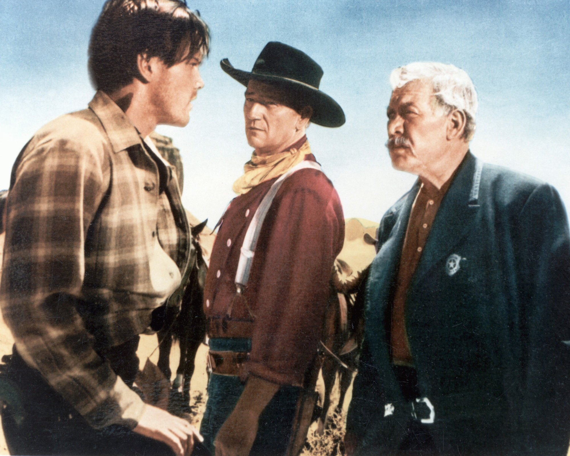 'The Searchers' Jeffrey Hunter as Martin Pawley, John Wayne as Ethan Edwards, and Ward Bond as Rev Capt. Samuel Johnston Clayton wearing Western clothes looking at each other with horses in the background