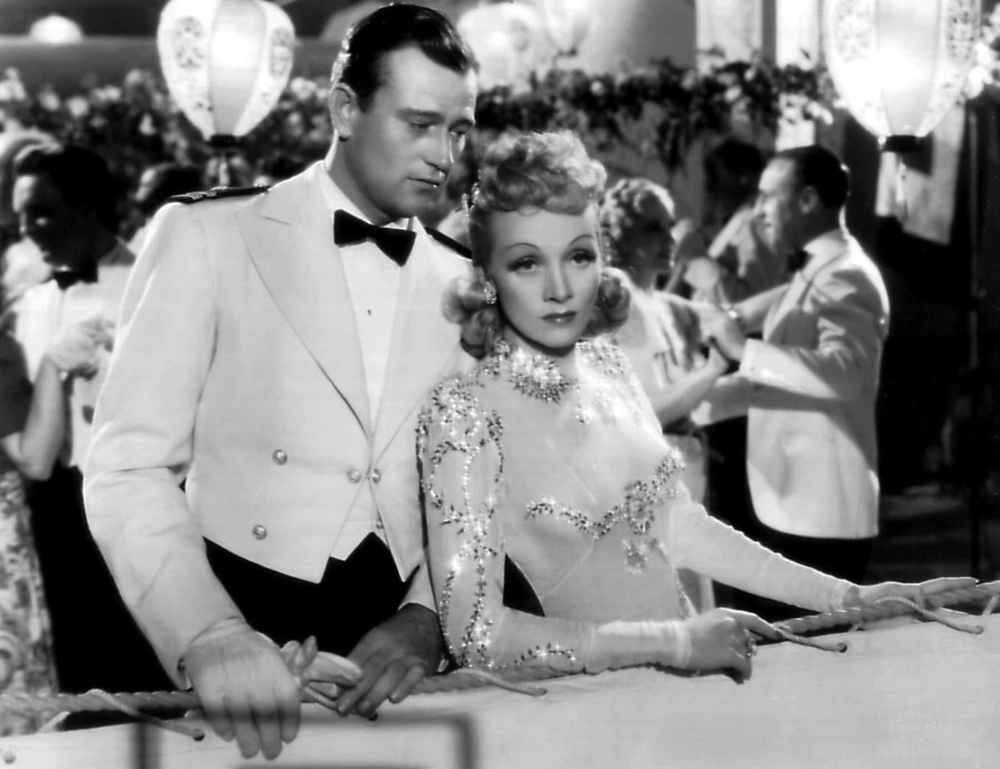 'The Seven Sinners' John Wayne as Dan and Marlene Dietrich as Bijou, who allegedly made him say the Pledge of Allegiance in between her thighs. They are holding onto a rope bannister, dressed in a tux and dress.