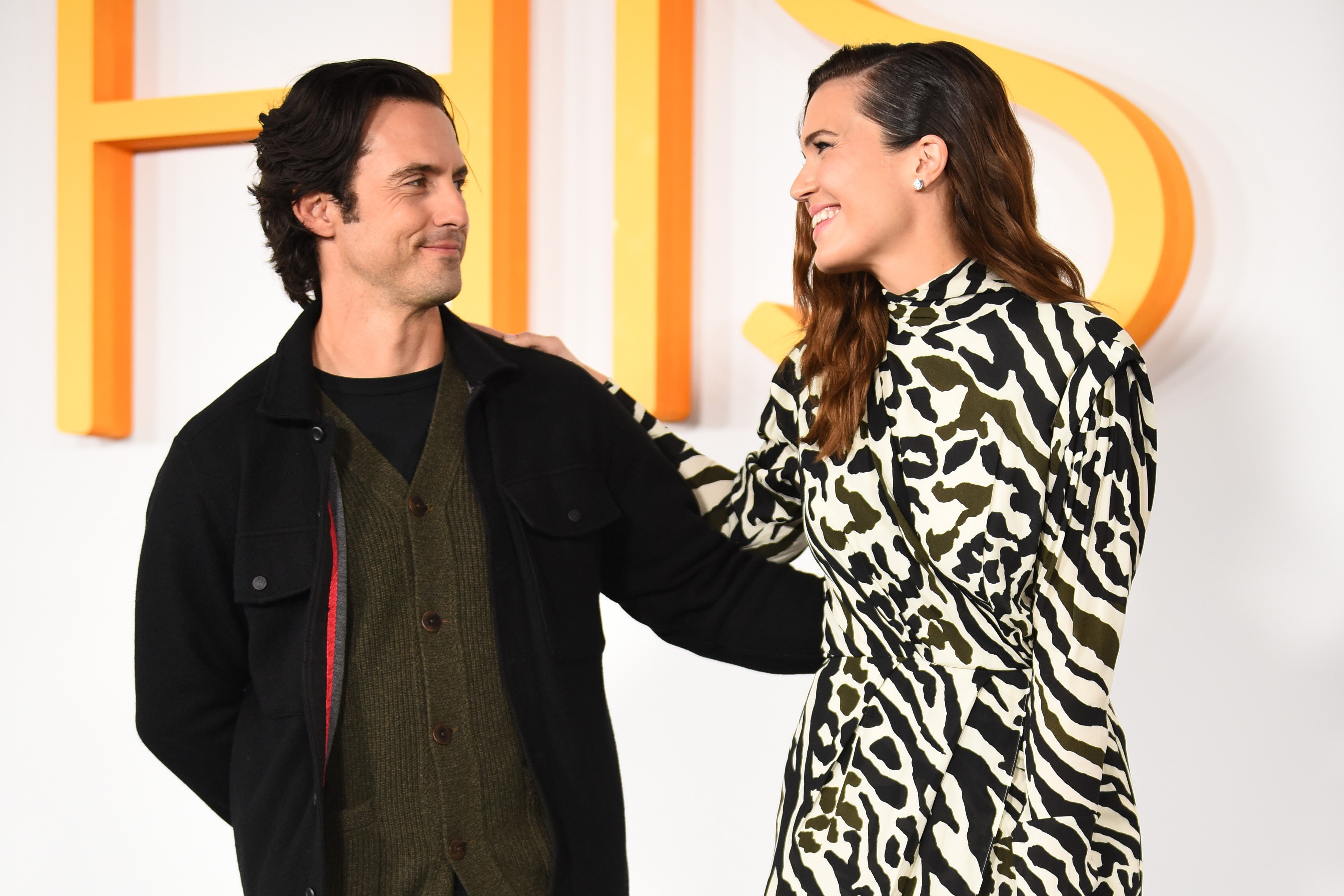 'This Is Us' stars Milo Ventimiglia and Mandy Moore pose for pictures together on the red carpet. Ventimiglia wears a black jacket over a dark green cardigan over a black t-shirt. Moore wears a long-sleeved black and white zebra print dress.