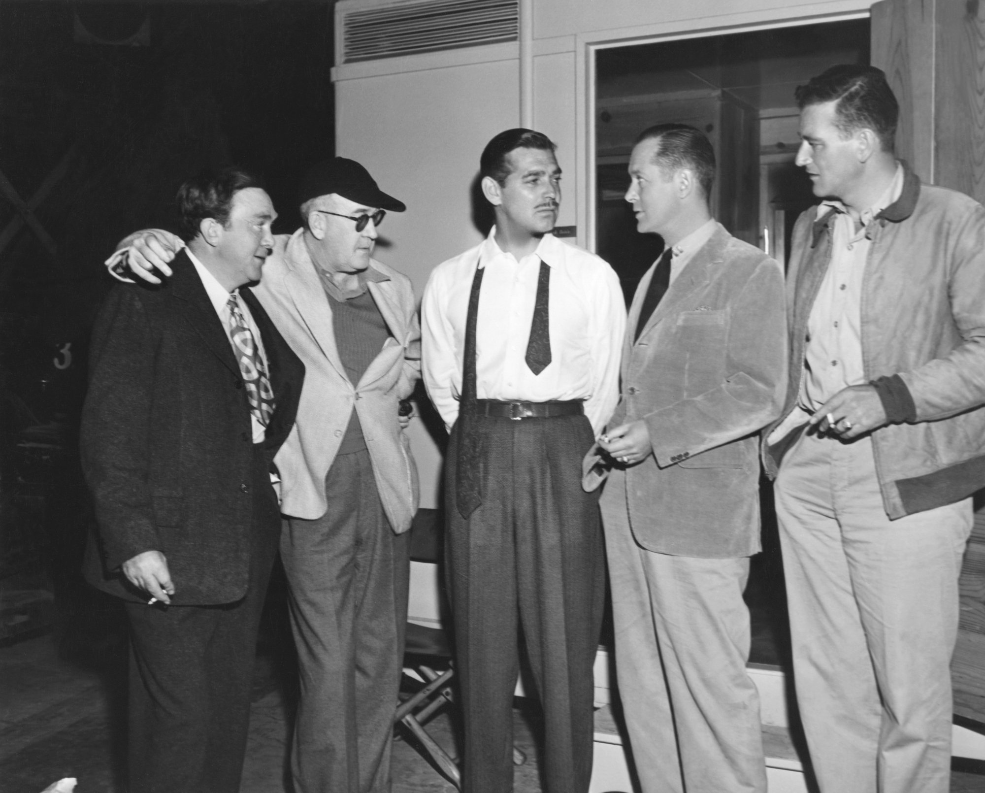 Thomas Mitchell, John Ford, Clark Gable, Robert Montgomery, and John Wayne standing around on the set wearing suits and smoking cigarettes