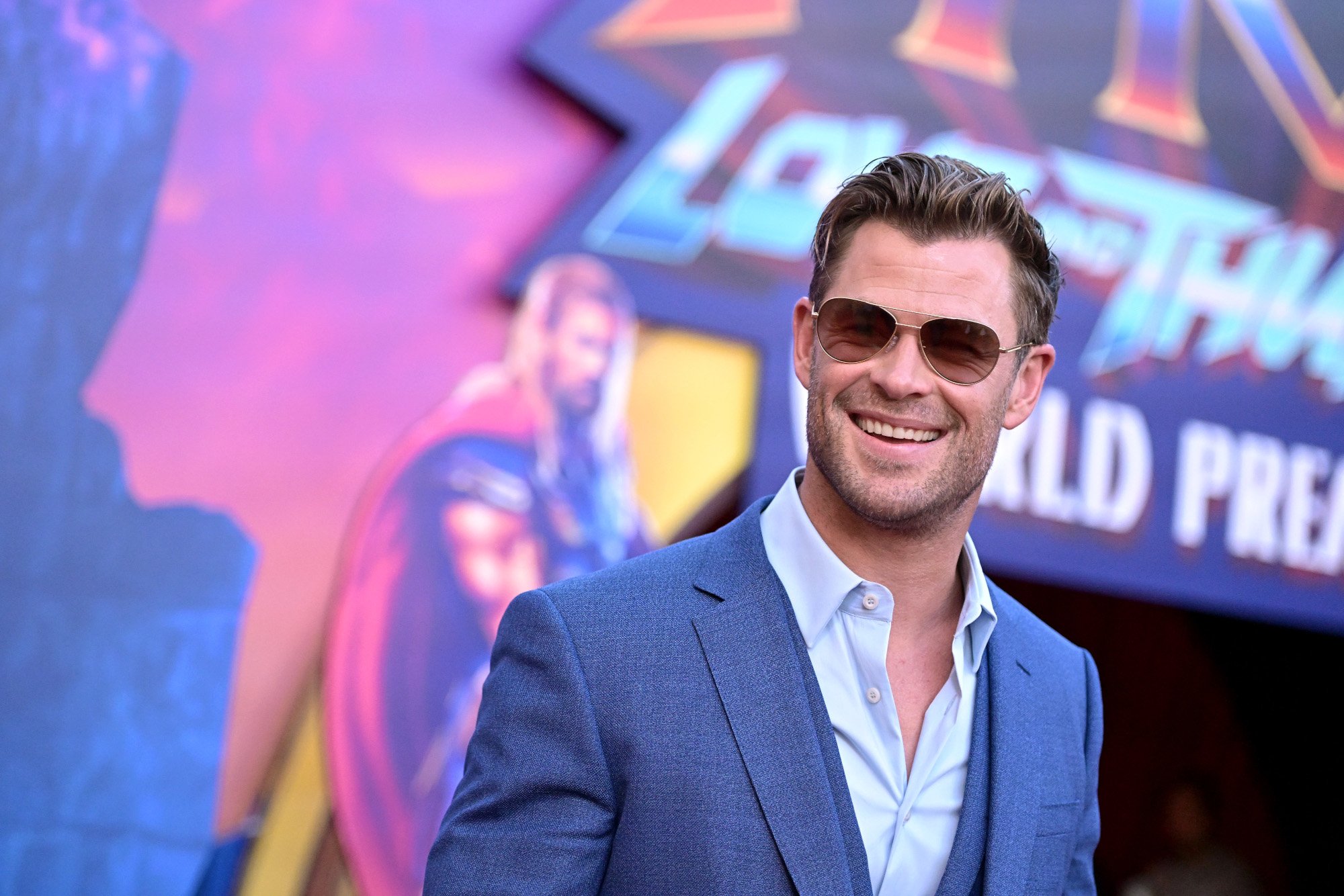Chris Hemsworth, who waited 10 years for his naked butt scene in 'Thor: Love and Thunder.' He's wearing sunglasses and smiling in this photo, with a blue button-up shirt and jacket on.