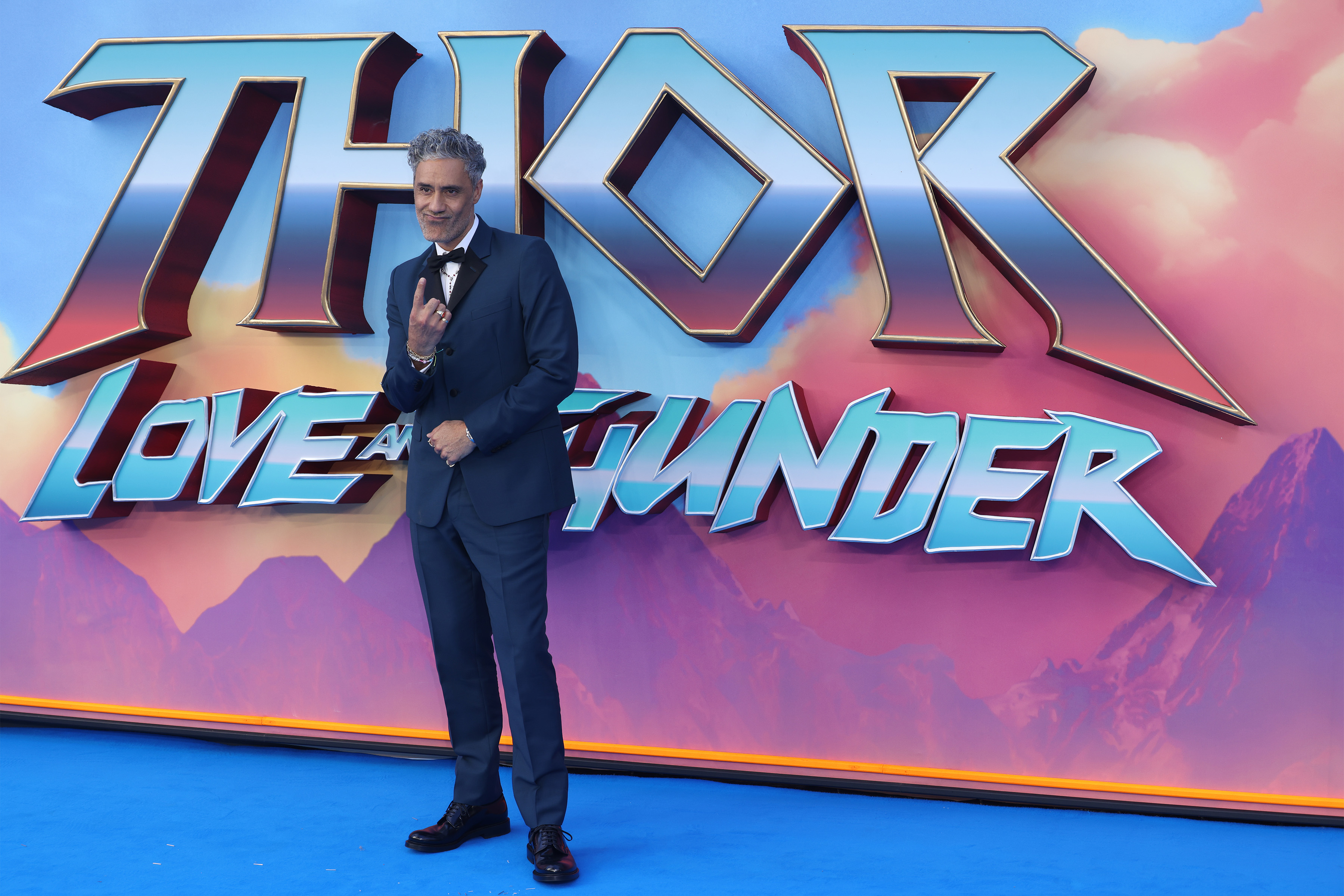 Taika Waititi, the director of 'Thor: Love and Thunder,' wears a dark blue suit while standing in front of a sign for the movie.