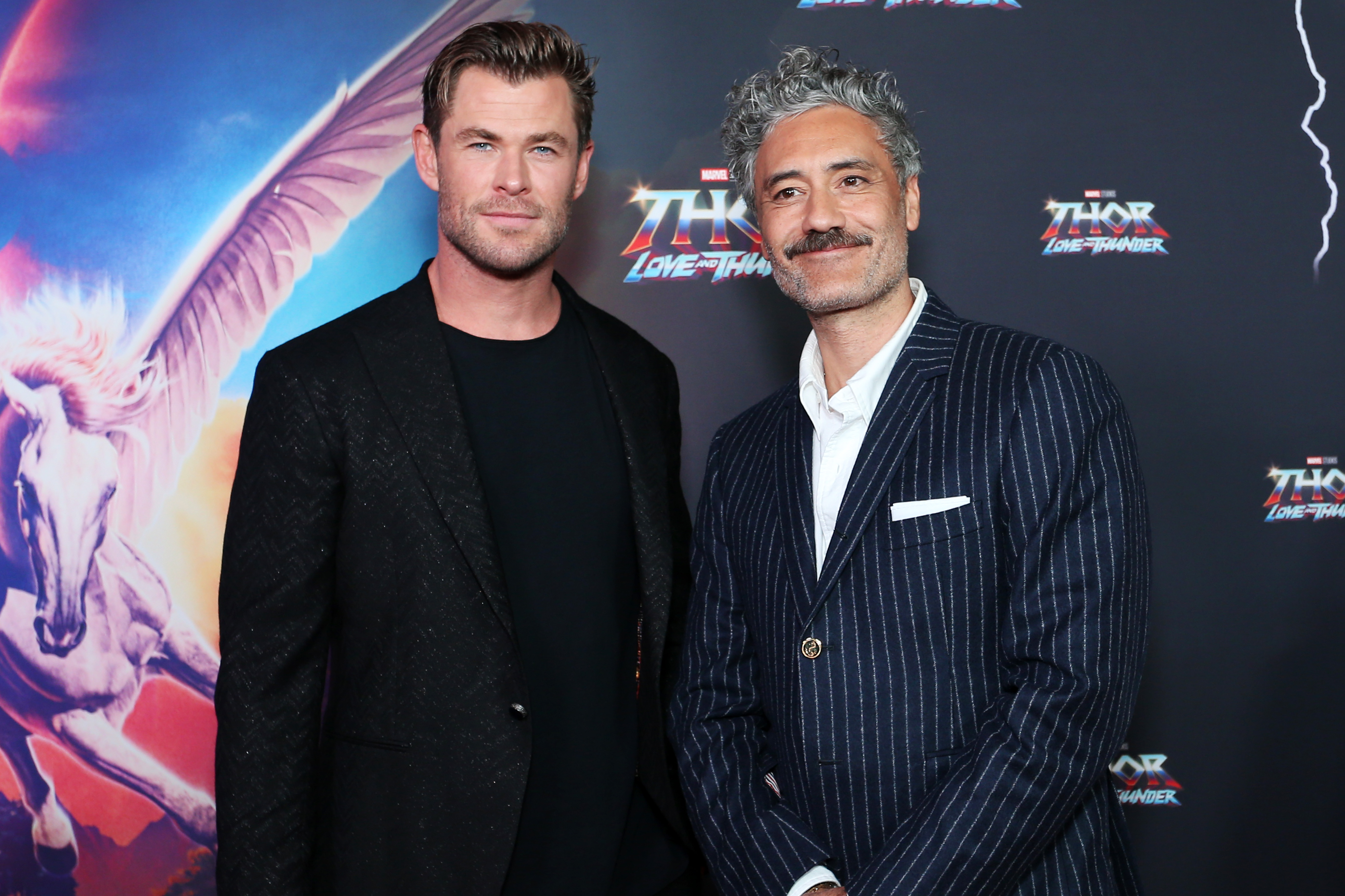 Chris Hemsworth and Taika Waititi, who star in Marvel's 'Thor: Love and Thunder,' pose for pictures on the red carpet. Hemsworth wears a black suit over a black shirt. Waititi wears a black suit with thin white vertical stripes over a white button-up shirt.