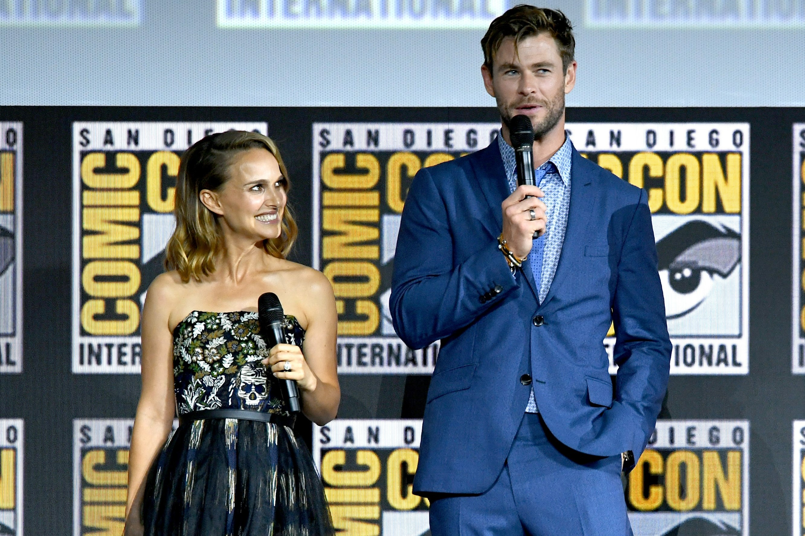 Natalie Portman and Chris Hemsworth, who star in 'Thor: Love and Thunder,' speak onstage at San Diego Comic-Con. Portman wears a black strapless dress with white and green leaves. Hemsworth wears a blue suit.