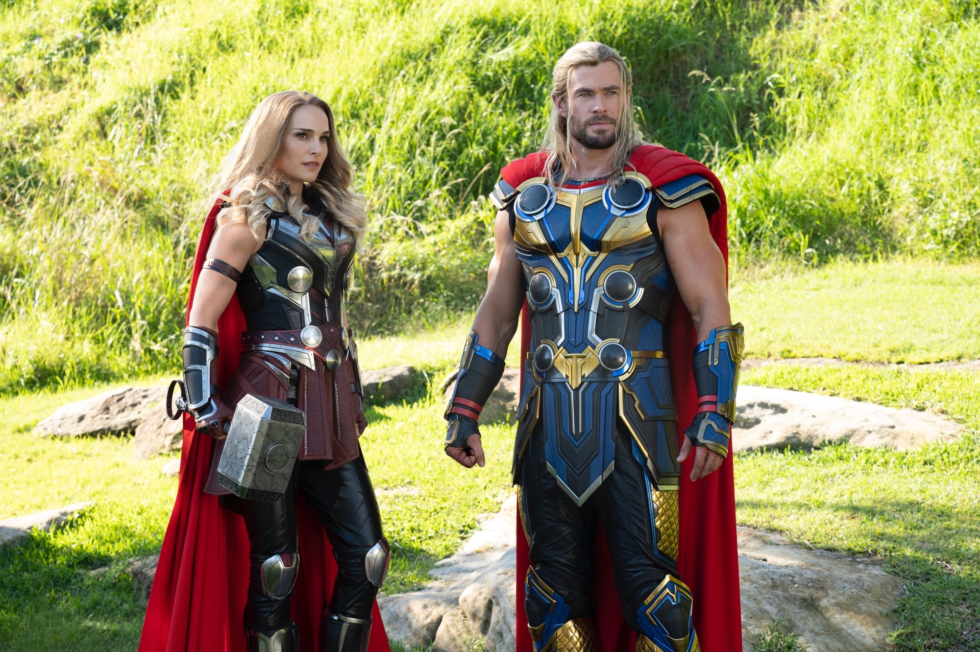 'Thor: Love and Thunder' Natalie Portman as Mighty Thor and Chris Hemsworth as Thor standing next to each other in their costumes in front of grassy hill