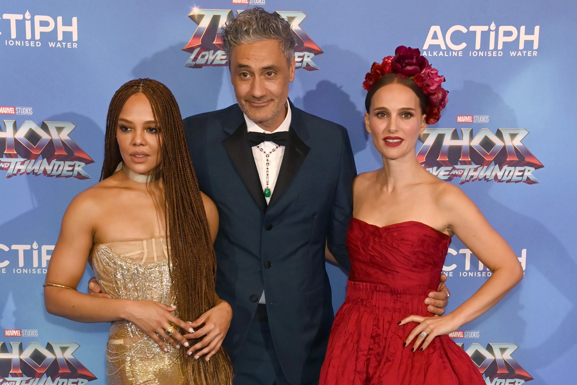 'Thor: Love and Thunder' Tessa Thompson, Taika Waititi, and Natalie Portman posing in front of a step and repeat for the movie. Thompson and Portman wear dresses and Waititi wears a tux.