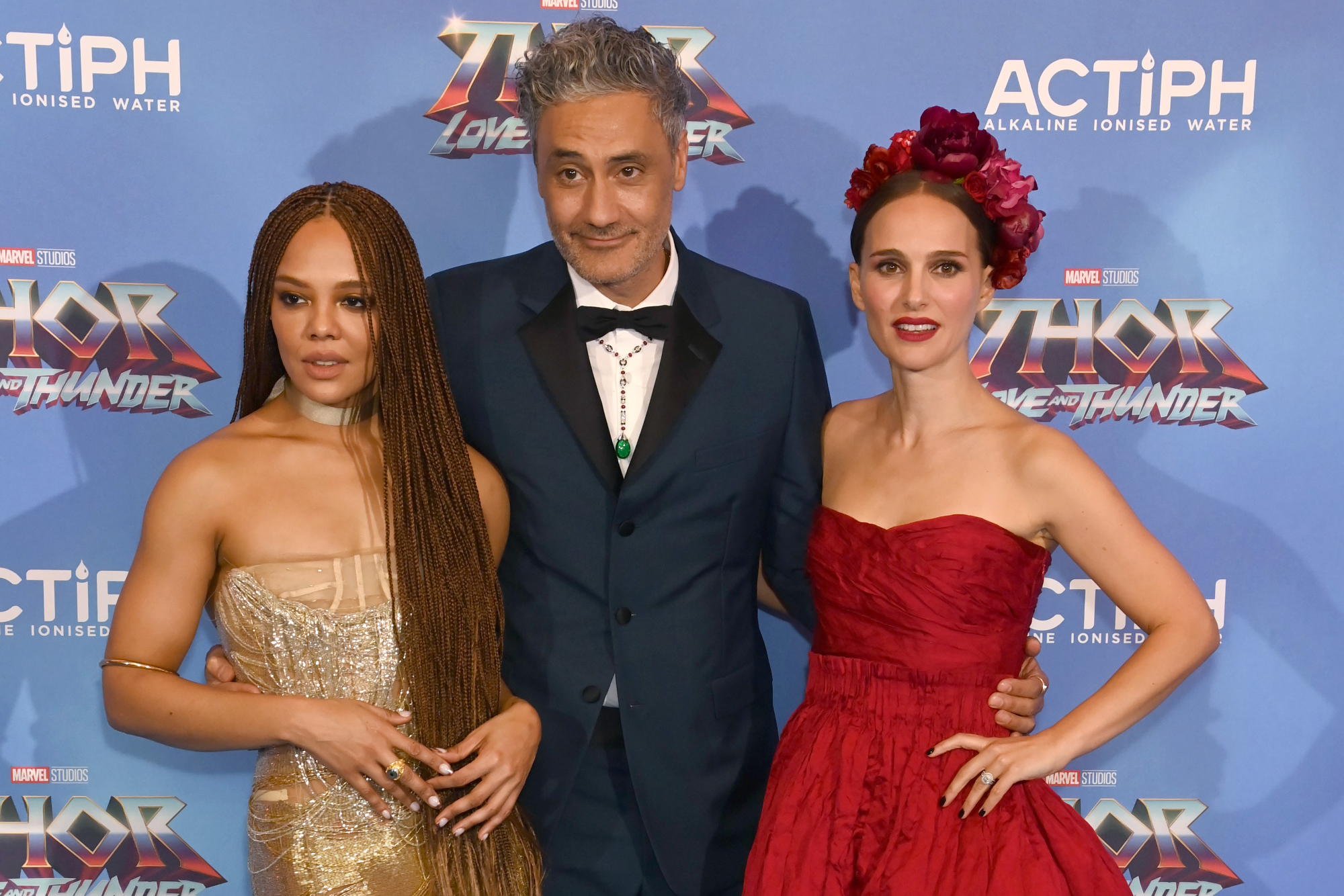 'Thor: Love and Thunder' stars Tessa Thompson, Taika Waititi, and Natalie Portman posing in front of a step and repeat for the movie. Thompson and Portman wear dresses, and Waititi wears a tux.