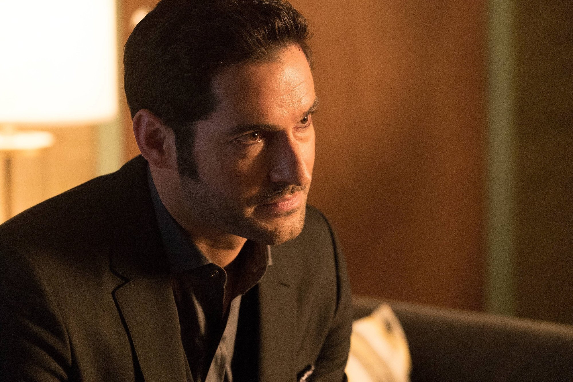 Tom Ellis, who 'Fantastic Four' fans want in the cast as Doctor Doom. He's wearing a black suit and looking intensely in front of him with a lamp in the background.