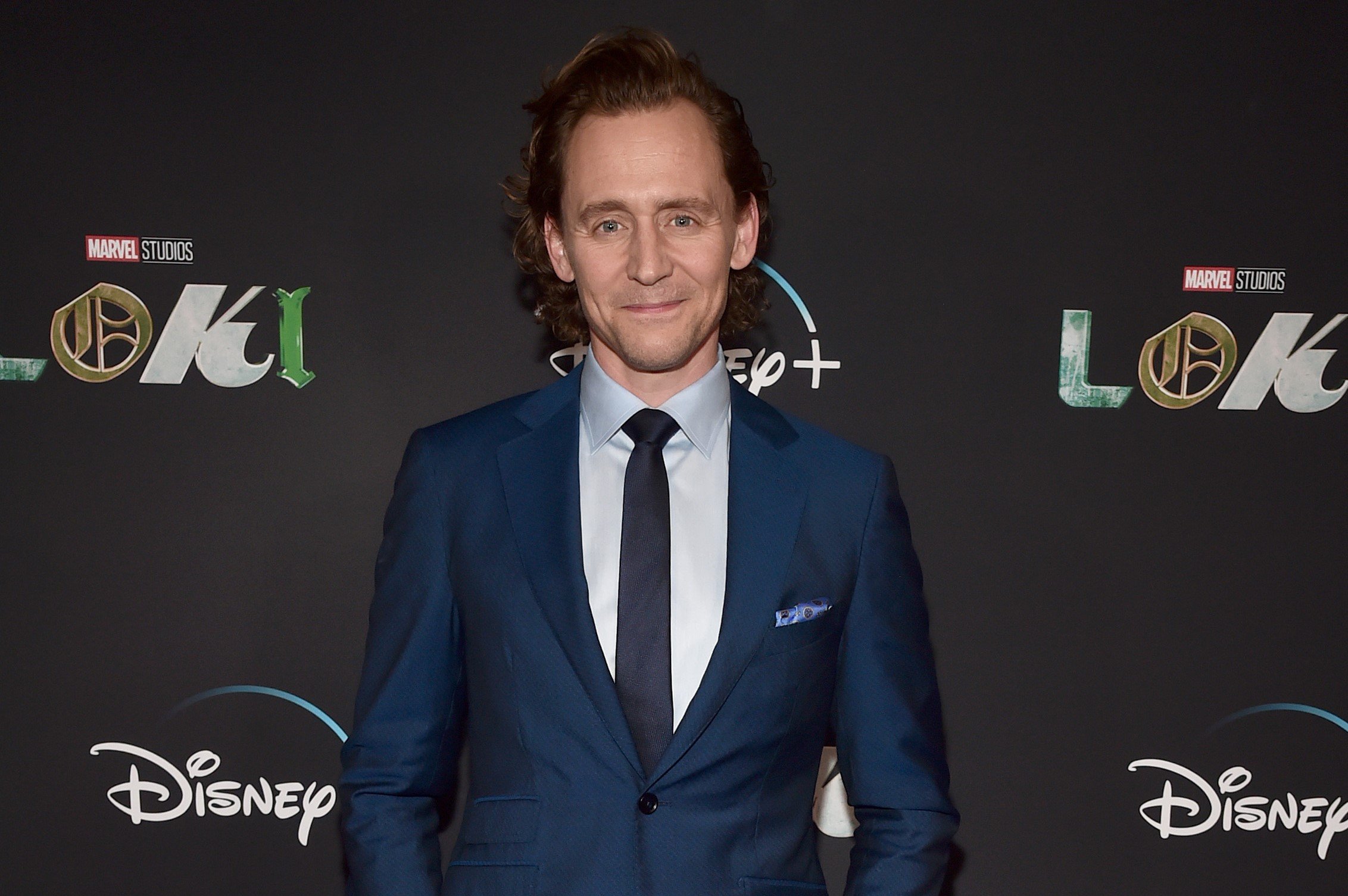 Tom Hiddleston, who played Loki in the 'Thor' movies, wears a dark blue suit over a light blue button-up shirt and dark blue tie.