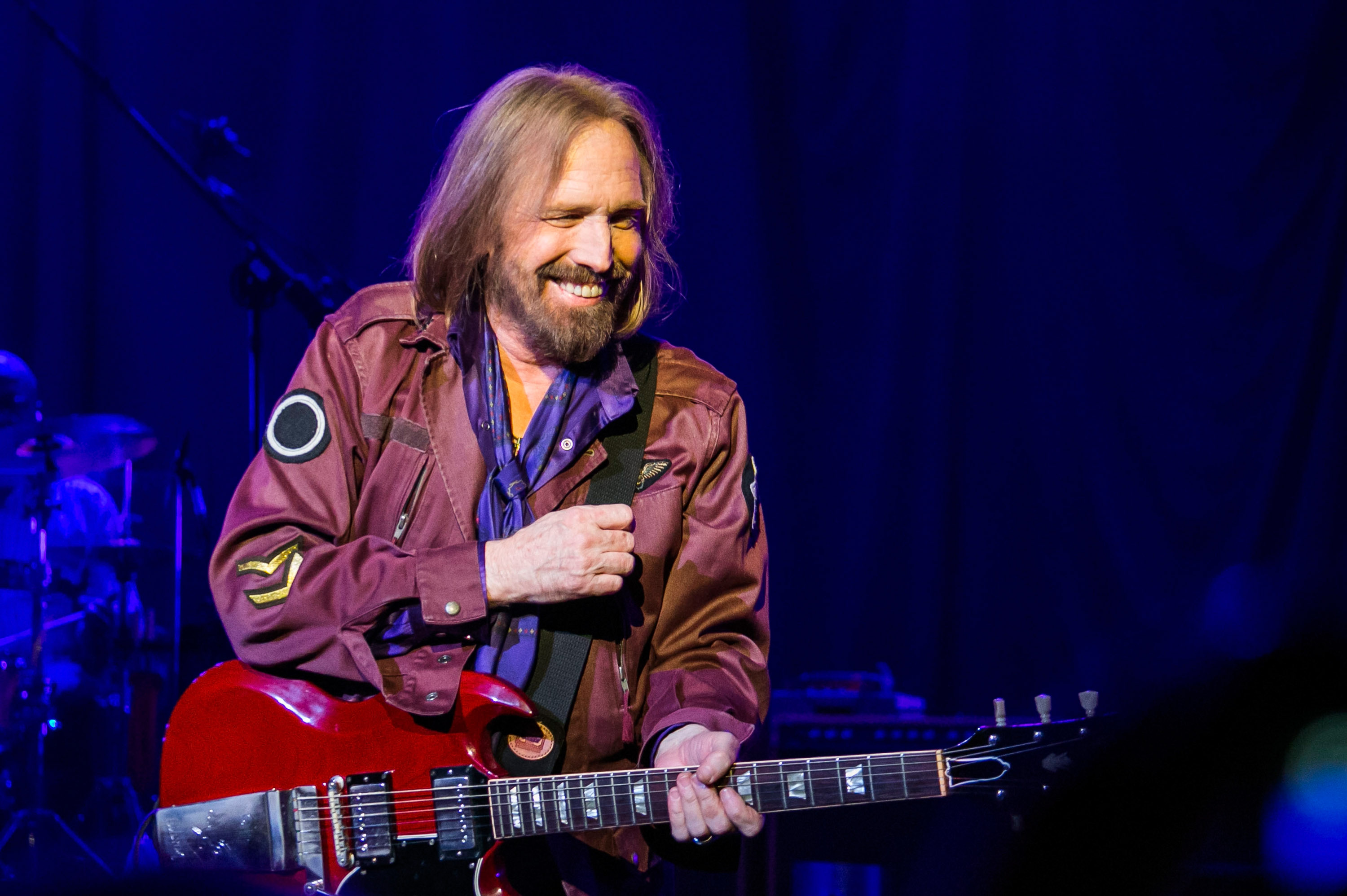 Tom Petty stands onstage and plays a red guitar.