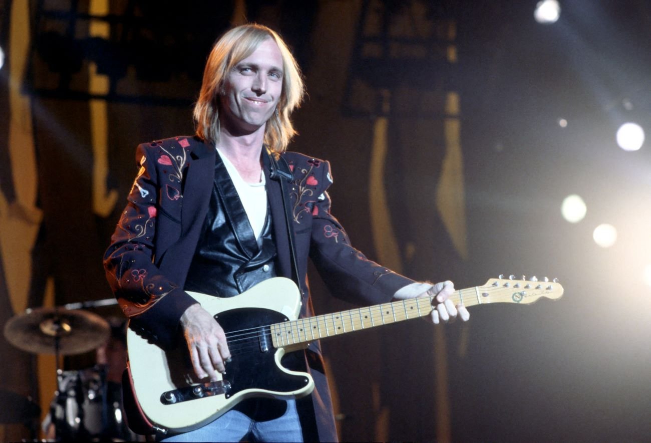 Tom Petty wears a black embroidered jacket and holds a white guitar.
