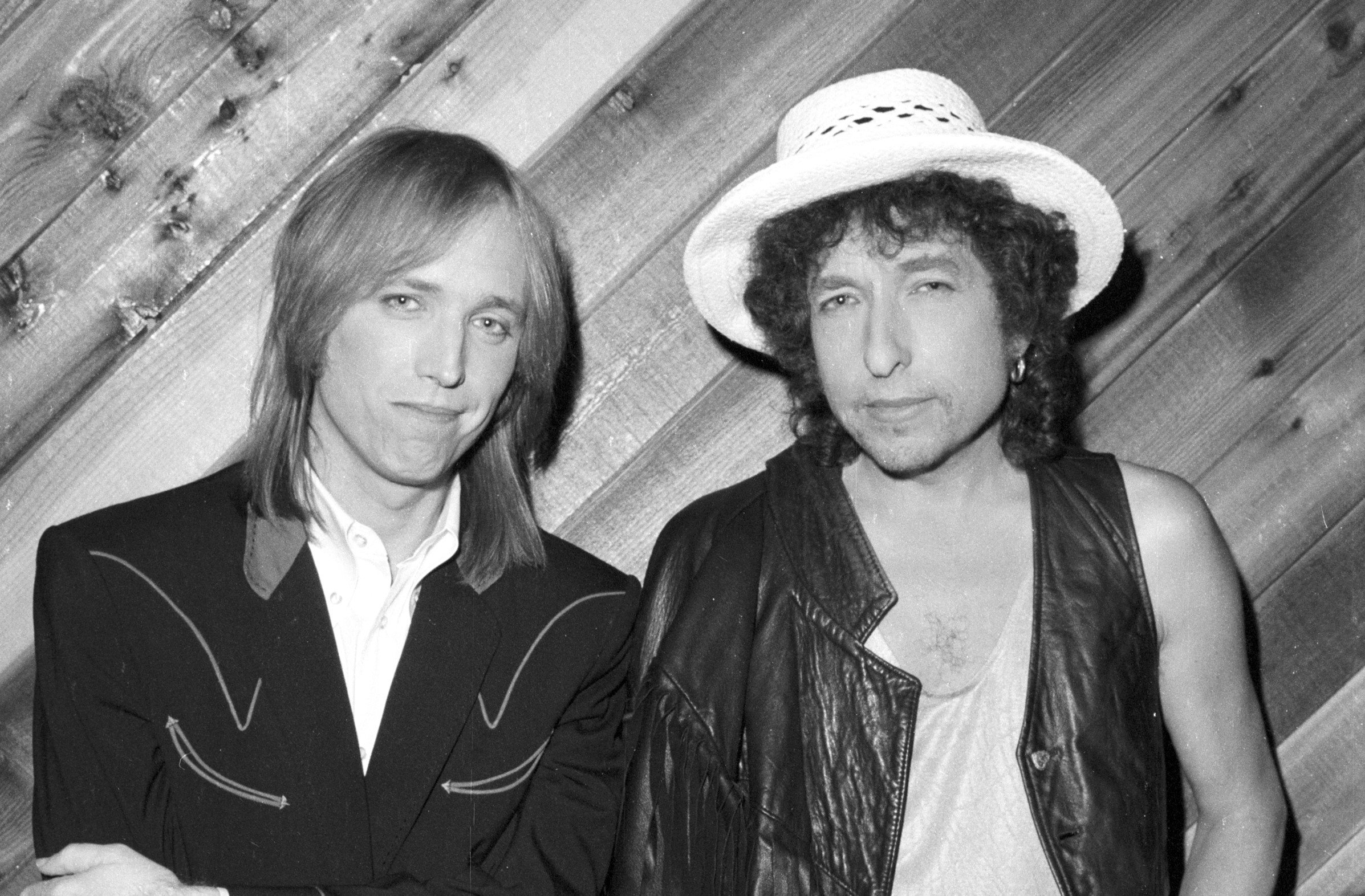 A black and white picture of Tom Petty and Bob Dylan standing in front of a wood paneled wall. 