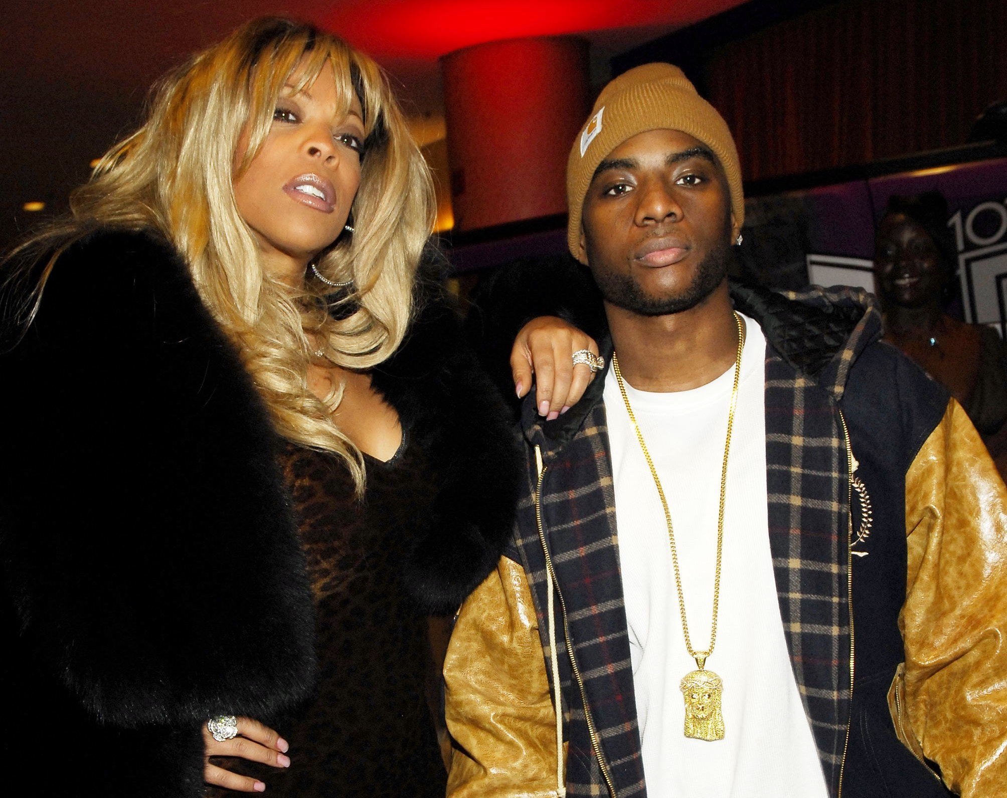 Wendy Williams wearing a black coat posing with her arm on Charlamagne tha God’s shoulder.
