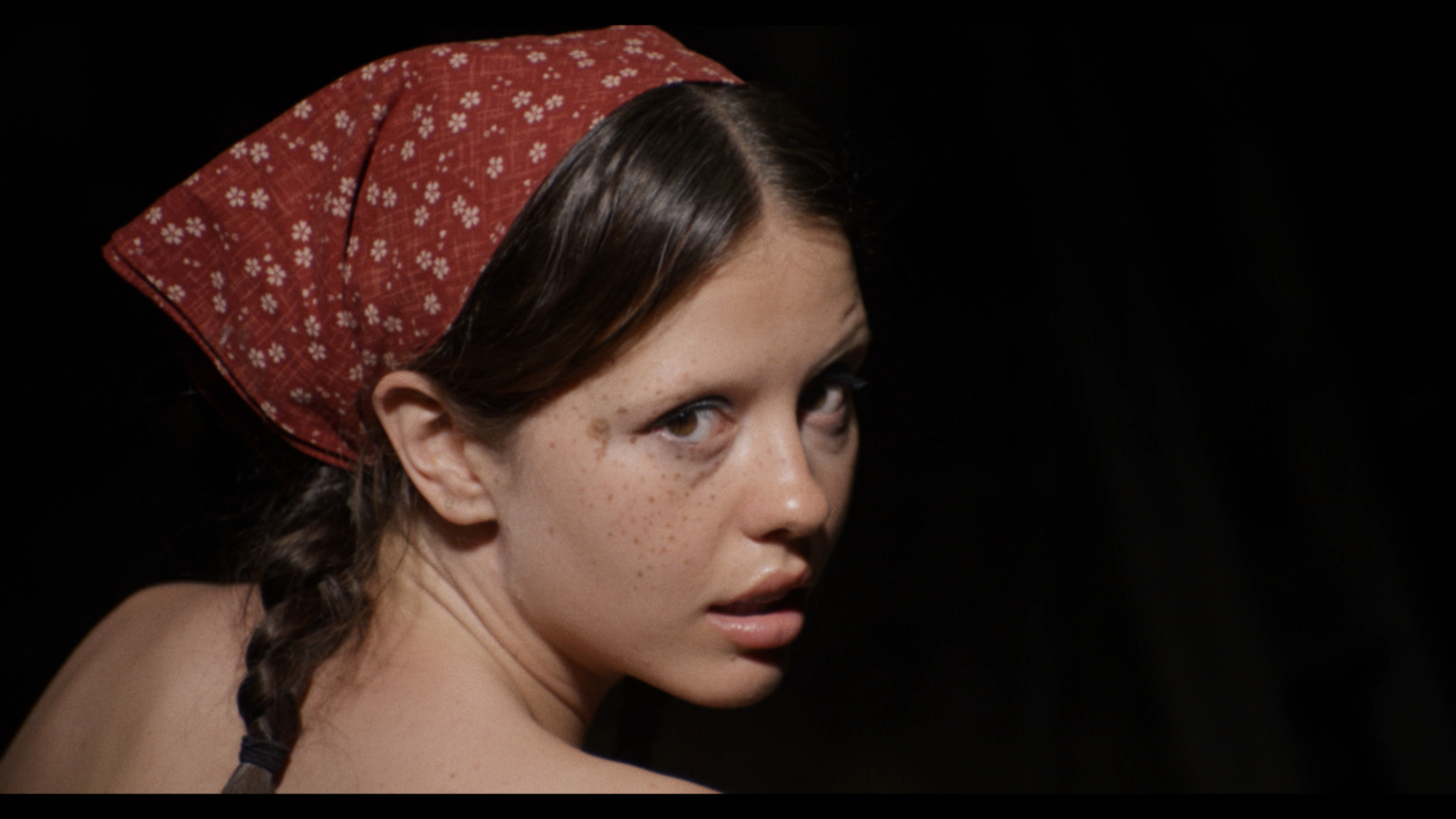 'X' Mia Goth as Maxine, who also stars as Pearl, looking over her shoulder wearing a bandana in her hair with her hair braided.