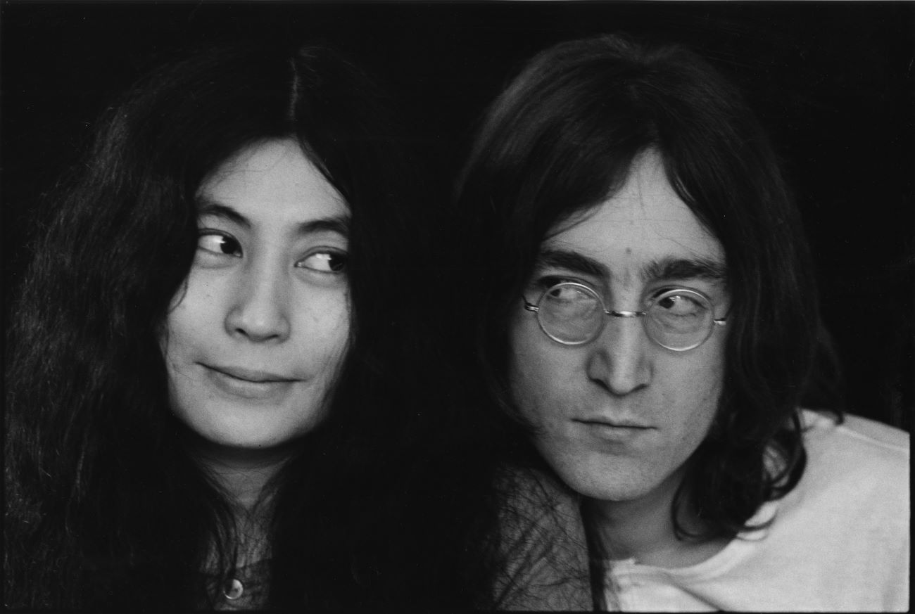 A black and white photo of Yoko Ono and John Lennon glancing sideways at each other.