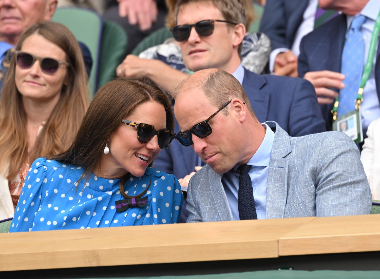 Prince William wears a light suit and Kate Middleton wears a blue polka dot dress as they lean in close to talk while watching Wimbledon tennis match