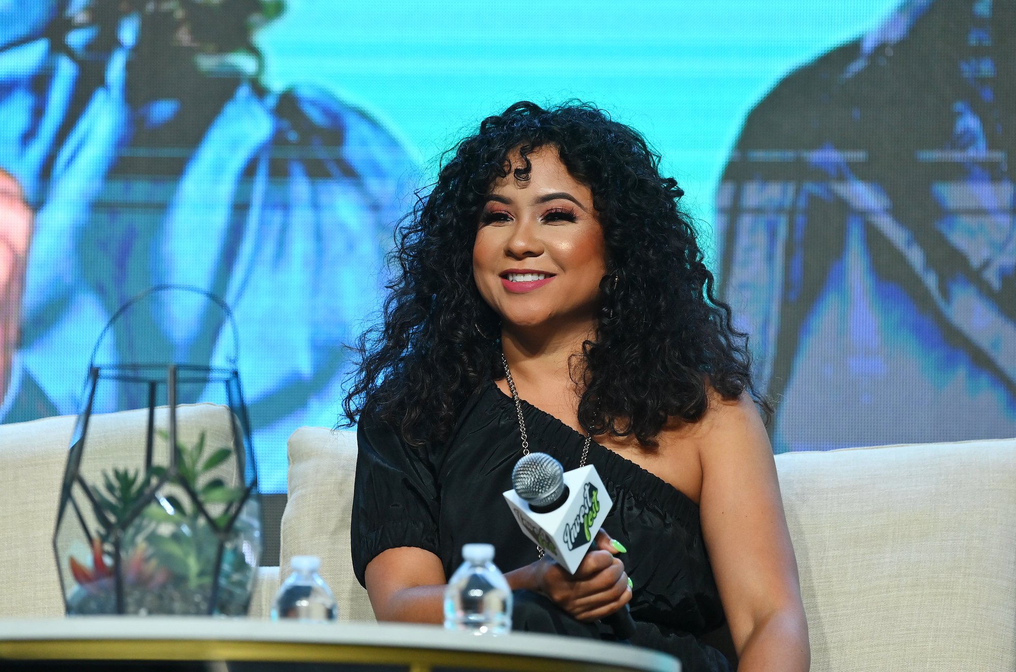 The Breakfast Club’ co-host Angela Yee smiling and holding a microphone while seated on stage.
