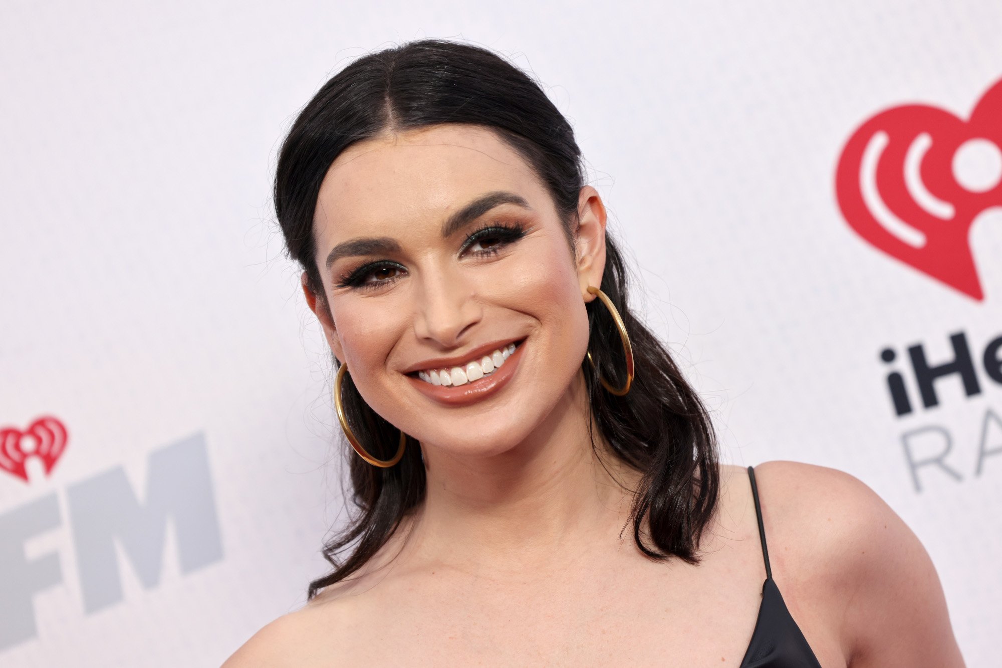 'Bachelor in Paradise' star Ashley Iaconetti, who recently revealed who she wants Rachel Recchia to end up with during 'The Bachelorette' 2022. In the photo, she's wearing a black dress, her hair is half-up and half-down, and she's standing in front of a wall with the iHeartRadio logo on it.