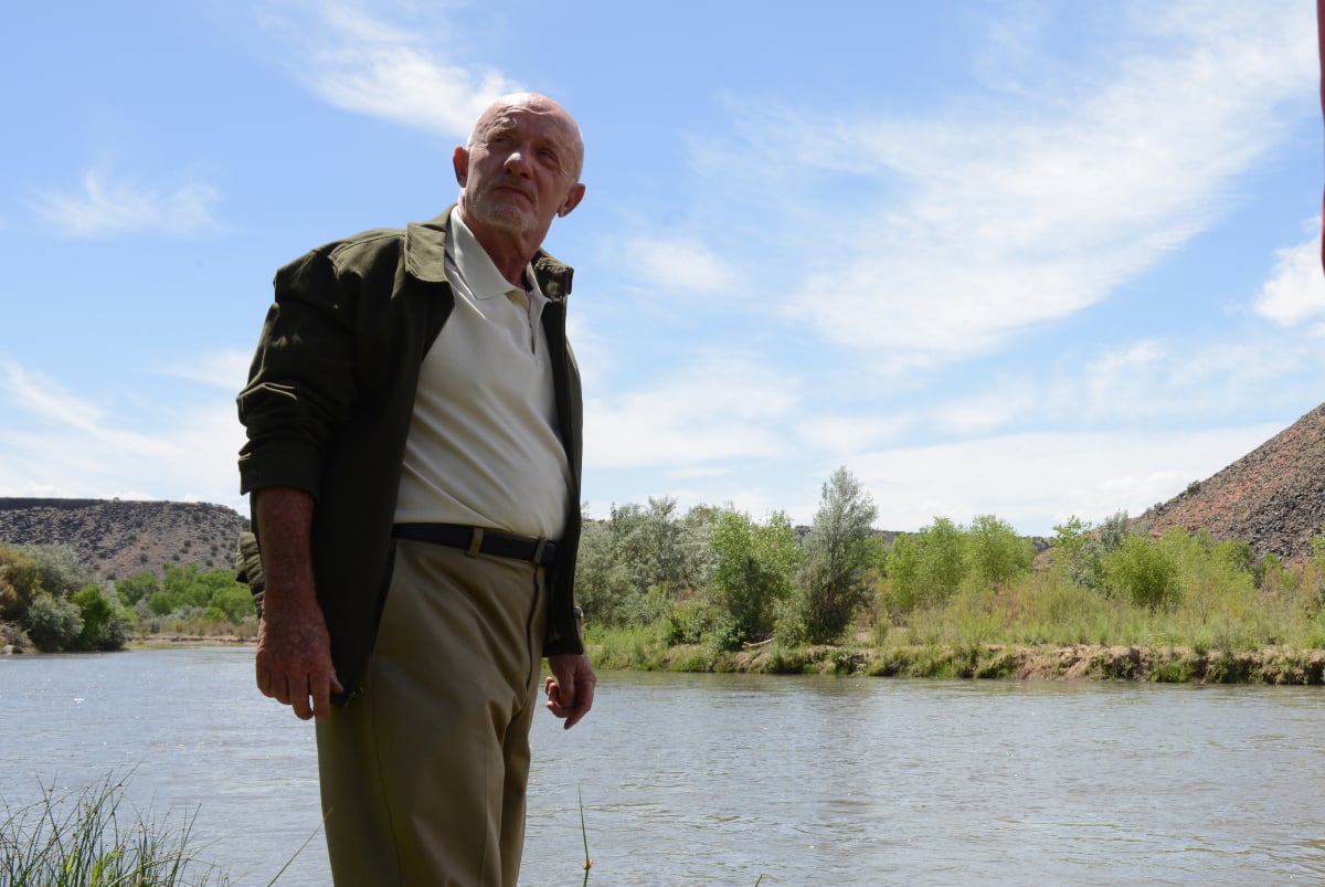 Jonathan Banks as Mike Ehrmantraut in Breaking Bad. Mike stands in front of a river wearing a khakis, a white polo shirt, and a jacket.