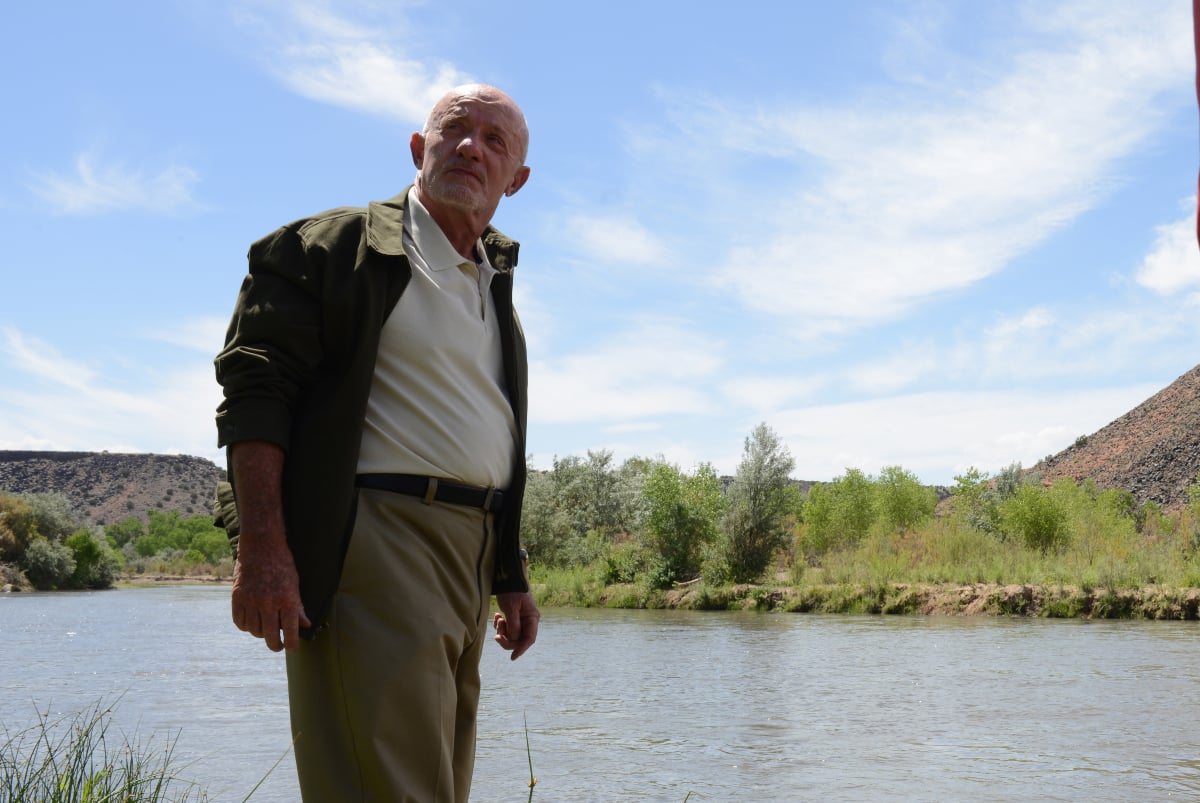 Jonathan Banks as Mike Ehrmantraut in Breaking Bad. Mike stands in front of a river wearing a khakis, a white polo shirt, and a jacket. 