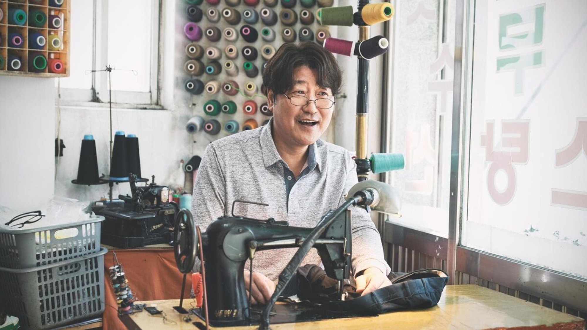 'Broker' Song Kang-ho as Sang-hyeon smiling while sitting behind a sewing machine while wearing glasses