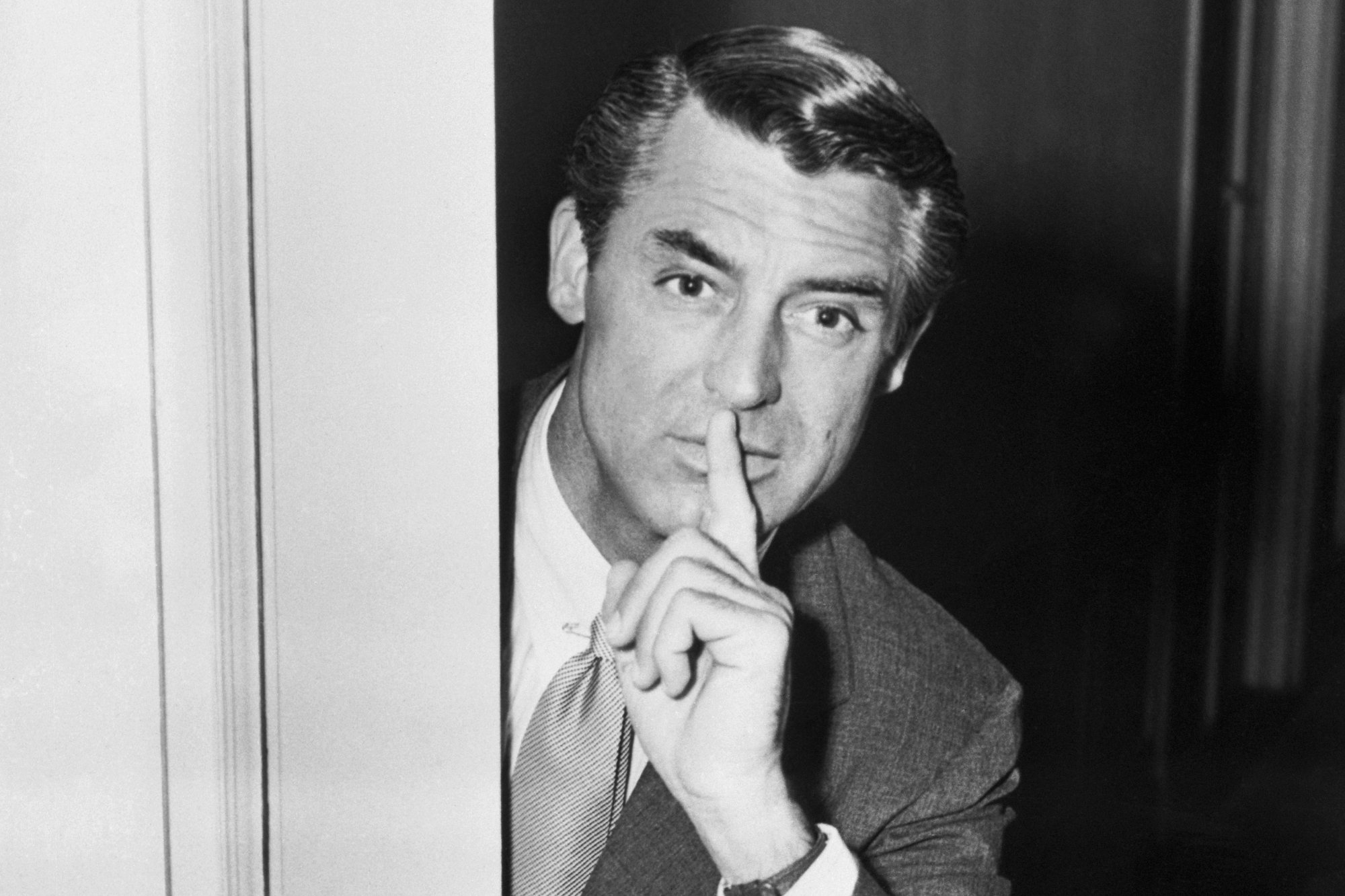 Cary Grant teasing movie project. He's holding a finger over his lips from behind a door. He's wearing a suit and tie.