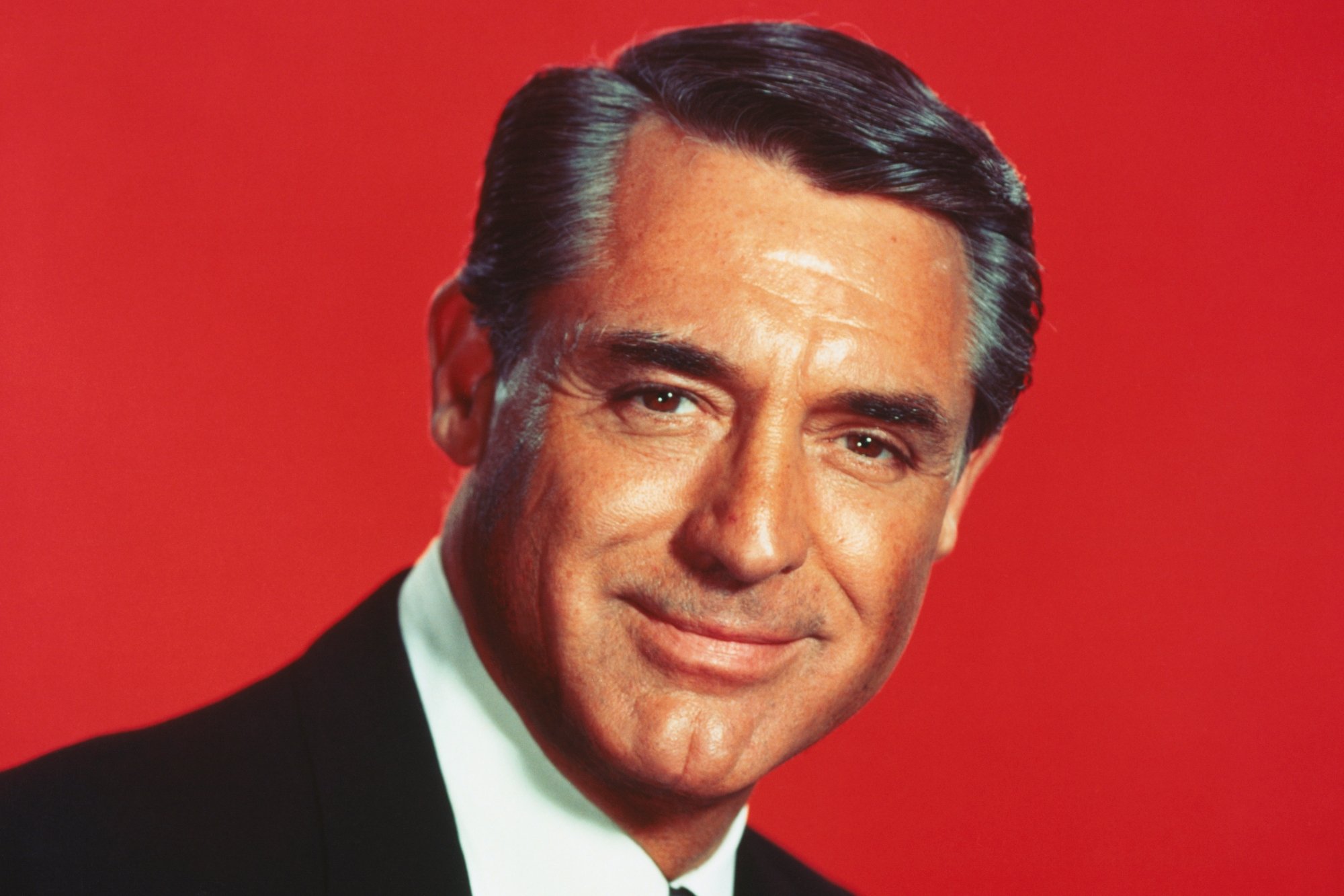 Cary Grant, who turned down 'Bridge on the River Kwai'. He's wearing a suit with a closed mouth smile in front of a solid red background.
