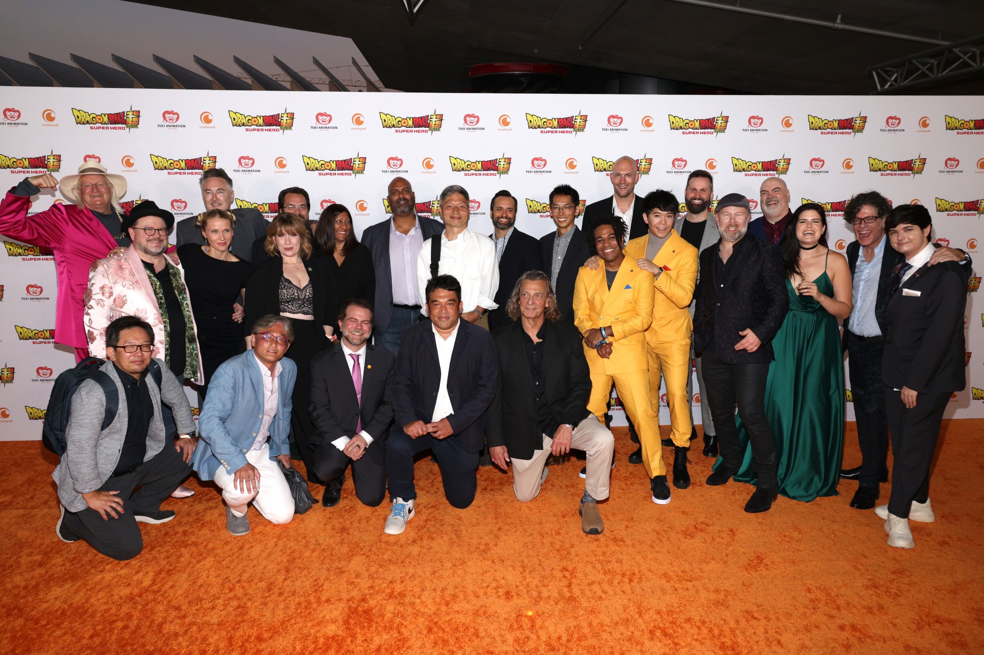 The cast of 'Dragon Ball Super: Super Hero' on the orange carpet for the film. They're all standing together in front of a wall with the movie's logo on it.