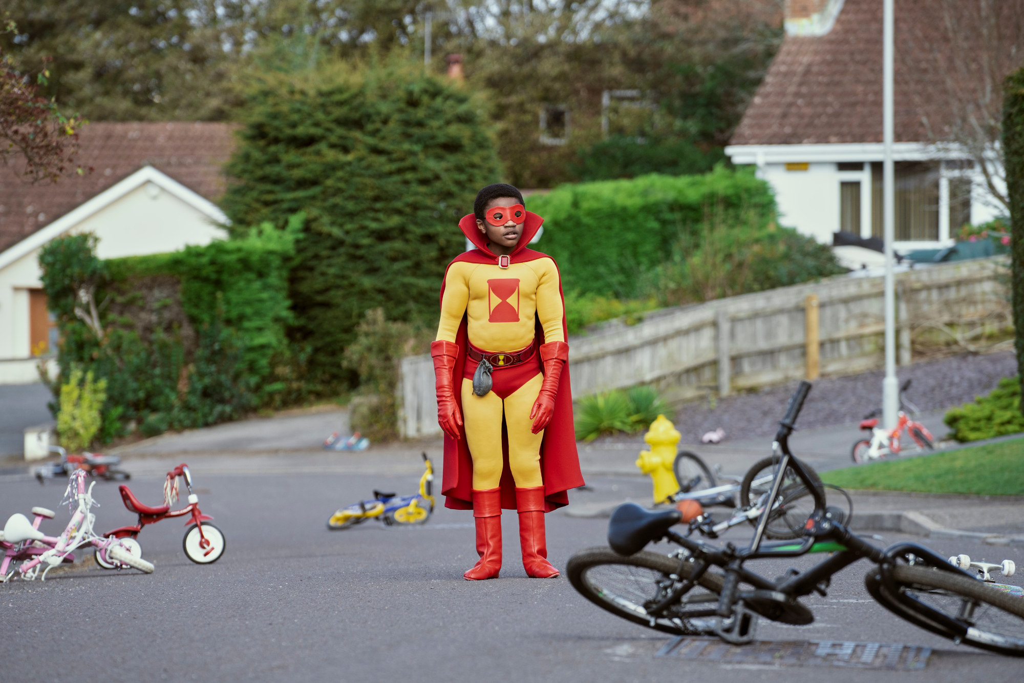 Eddie Karanja as Jed Walker in 'The Sandman.' He's wearing a yellow and red superhero suit and standing on a street with bikes tossed in it.