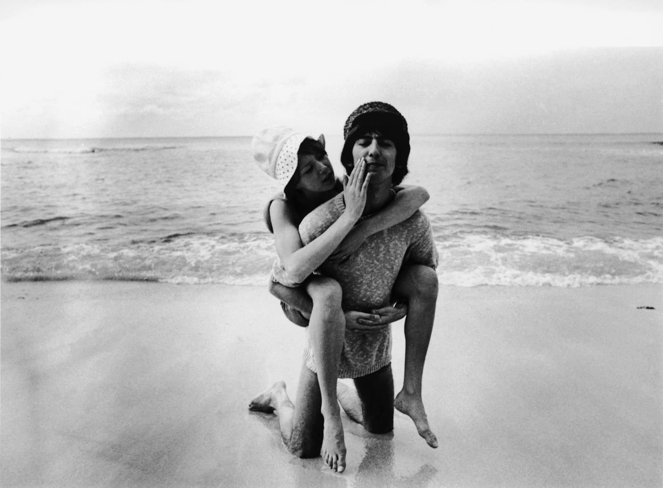 George Harrison kneels on a beach and Pattie Boyd sits on his back.