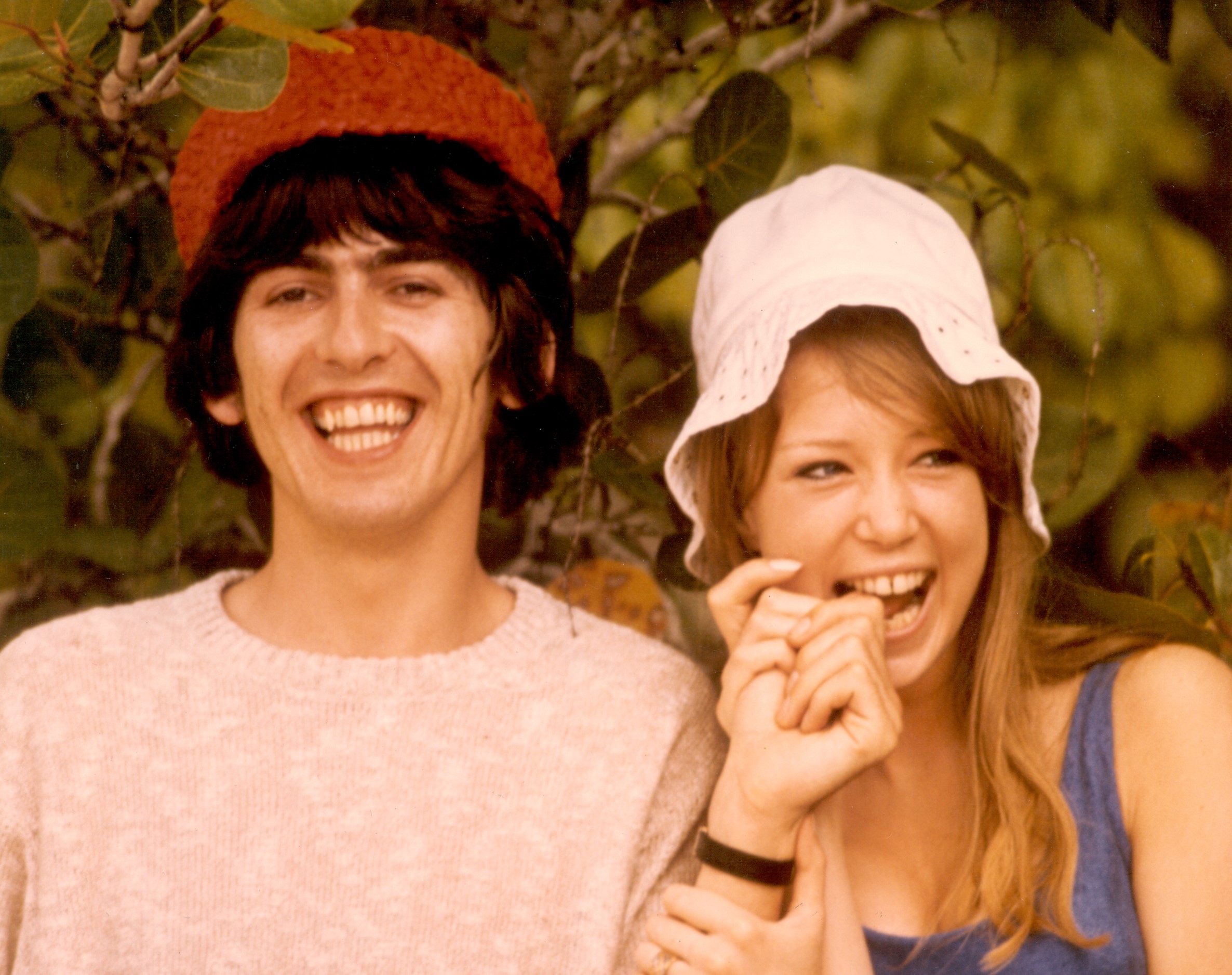 George Harrison and Pattie Boyd stand in front of a tree and wear hats. She bits his finger.