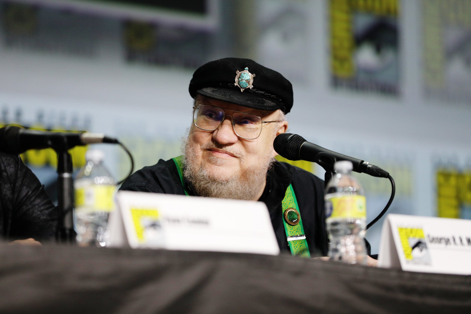'Game of Thrones' author George R.R. Martin, who sent signed copies of 'Fire & Blood' to the 'House of the Dragon' cast. In the photo, he's sitting down at a Comic-Con panel and wearing a hat. There's a microphone on the table in front of him.