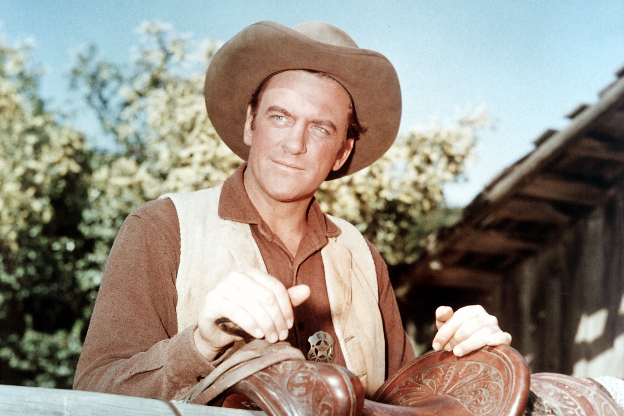 'Gunsmoke' James Arness as Matt Dillon with his hands on a horse saddle. He's wearing a cowboy hat and vest.