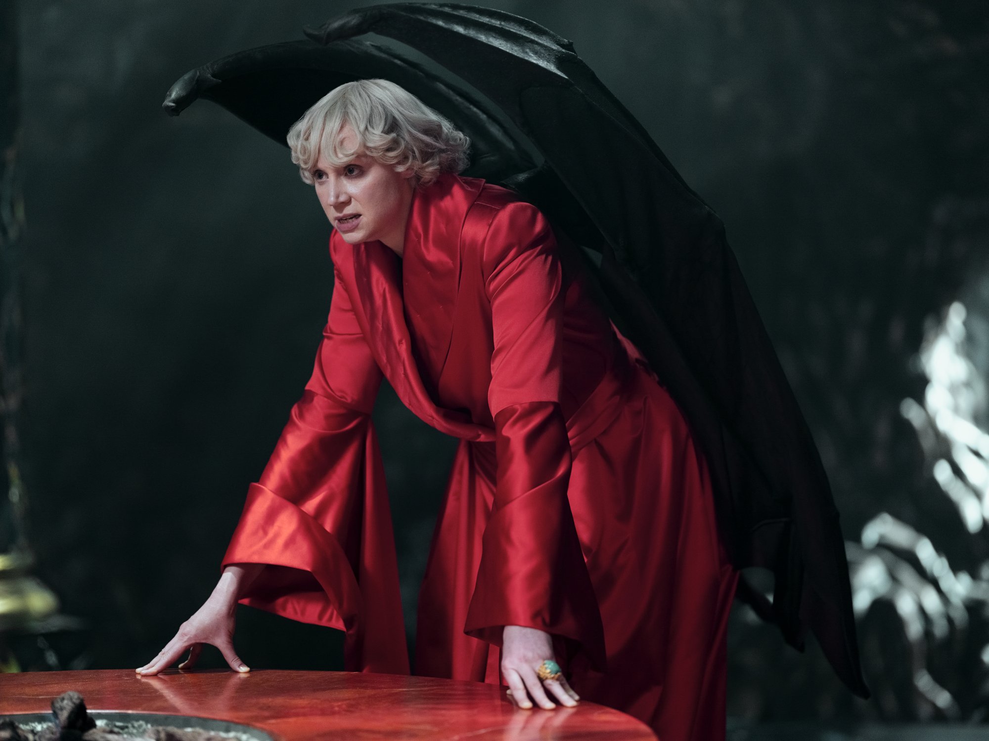 Actor Gwendoline Christie as Lucifer Morningstar in Netflix's 'The Sandman.' She's wearing all red, has black wings protruding from her back, and is leaning over a table.