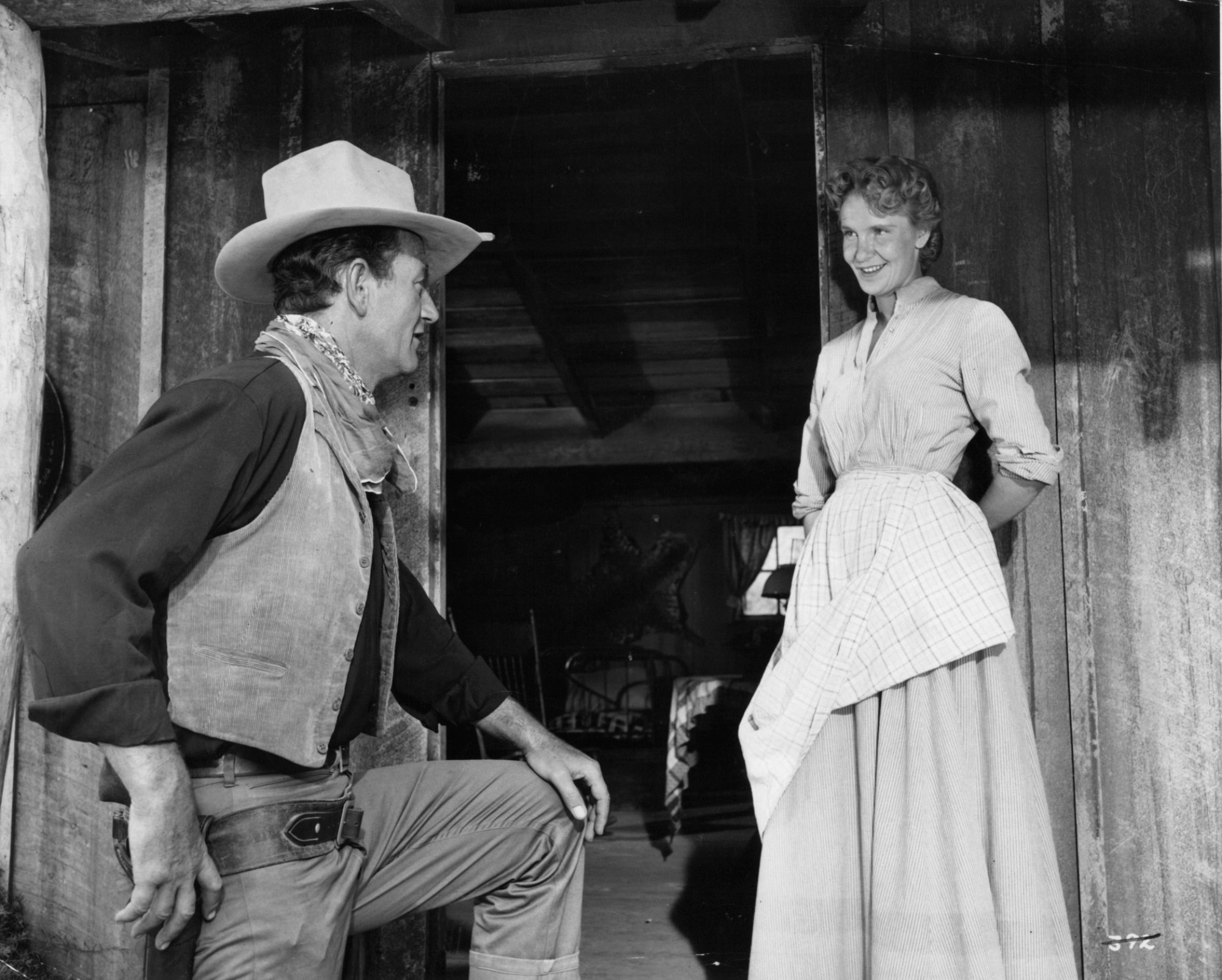 'Hondo' John Wayne as Hondo Lane and Geraldine Page as Angie Lowe. He's resting his leg on an upper stair, wearing a cowboy uniform looking at Page. She's smiling with her hands behind her back wearing an old-fashioned dress and apron.