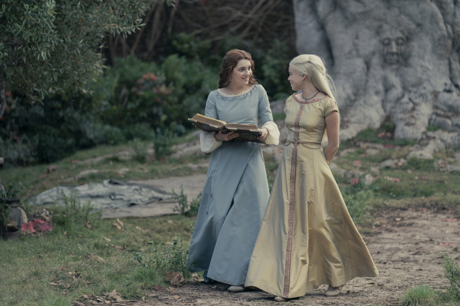 Emily Carey and Milly Alcock as Alicent Hightower and Rhaenyra Targaryen in 'House of the Dragon' Episode 1. They're wearing gowns and standing beneath a tree. Alicent is holding a large book in her hands.