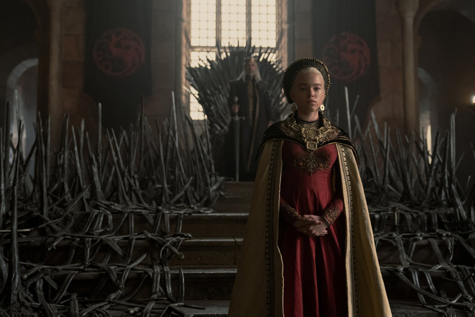 Paddy Considine and Milly Alcock as King Viserys Targaryen and Princess Rhaenyra in 'The House of the Dragon' for our review. King Viserys is sitting on the Iron Throne, and Princess Rhaenyra is standing in front of it. She's wearing a red and gold dress.