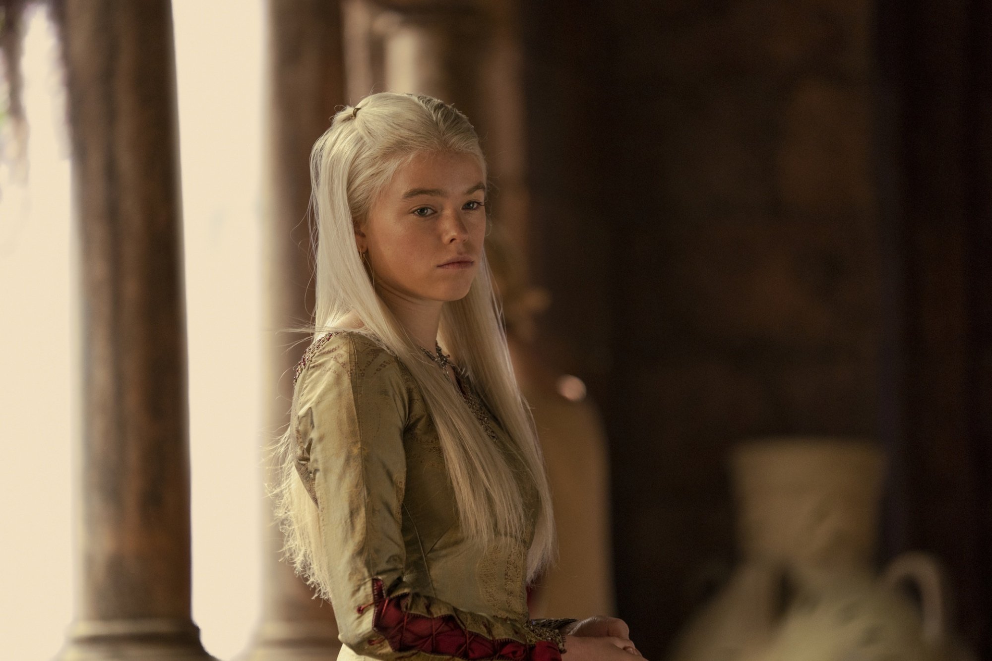 'House of the Dragon' opening theme introducing Milly Alcock as Young Princess Rhaenyra Targaryen wearing a gold dress with a serious look on her face. There's a large vase and a pillar in the background.